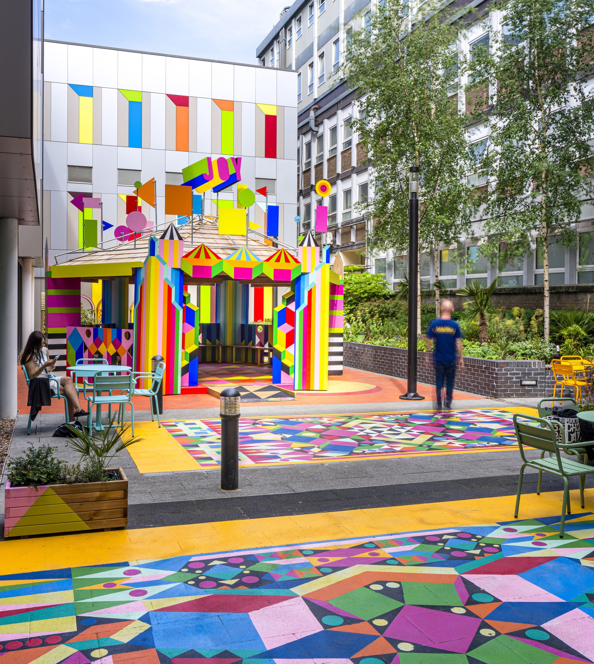 Outdoor pavilion with striped multicolor facade and matching ground graphics beside planted beds and seating