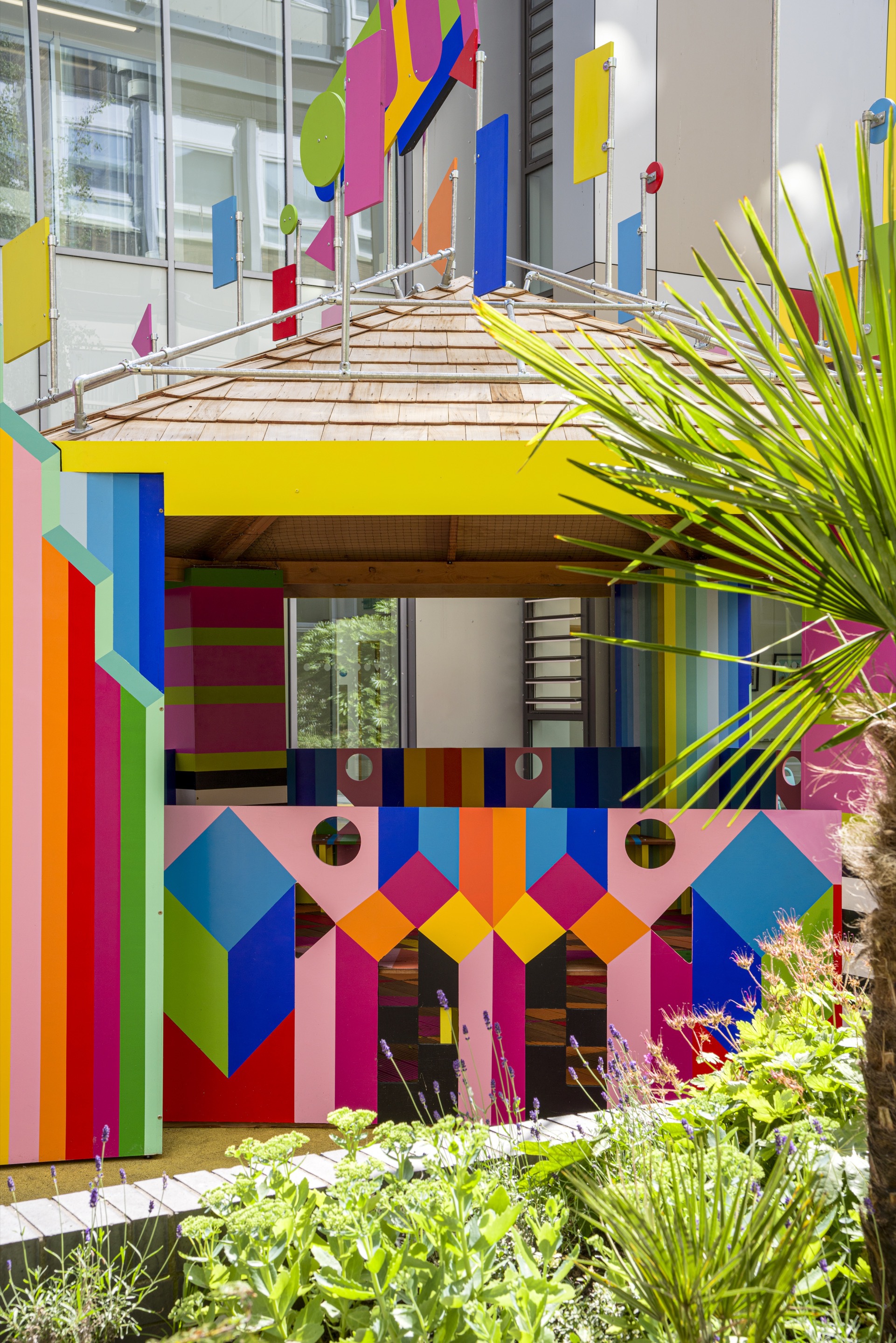 Striped timber roof canopy over multicolored pavilion facade framed by palm fronds and flowering plants