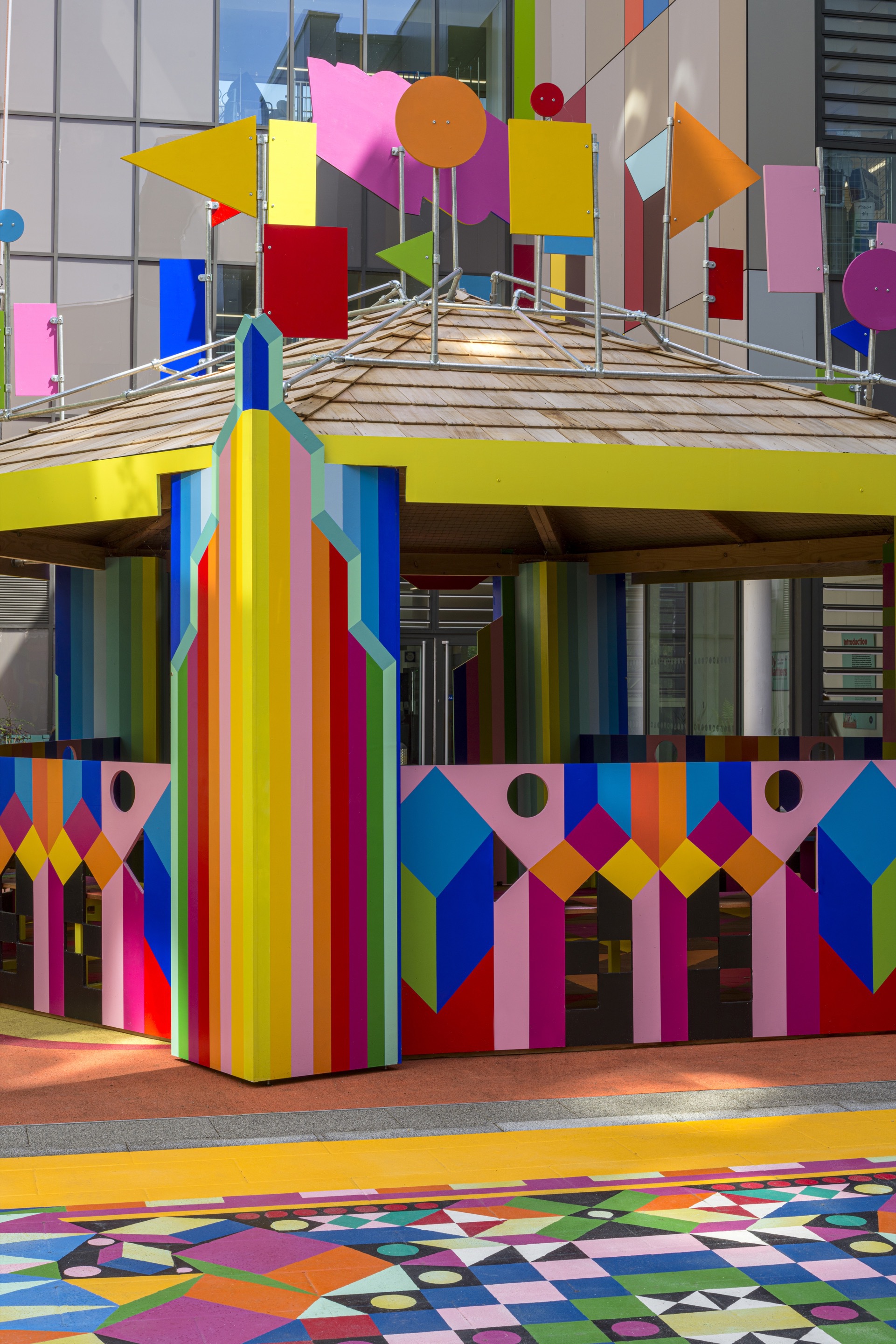 Pavilion structure with painted vertical stripe columns supporting a slatted timber roof under cloudy sky