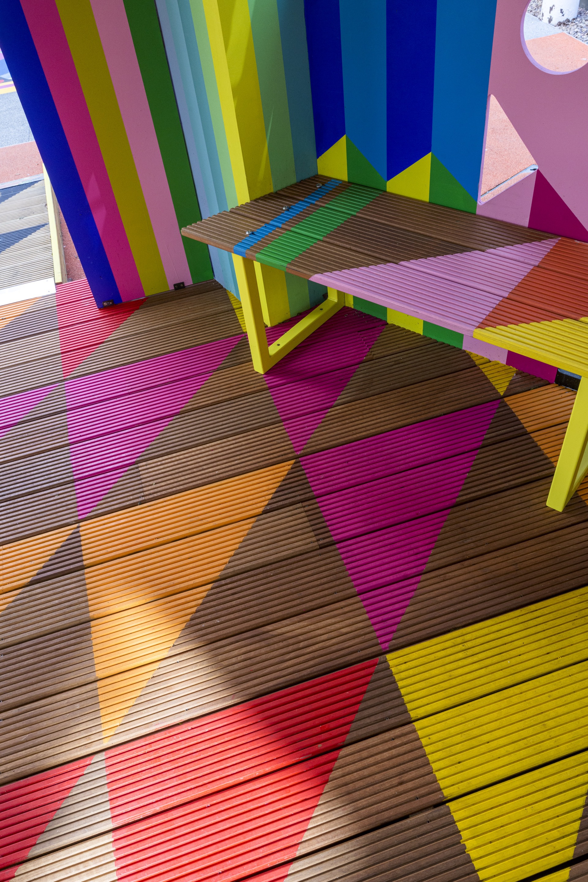 Overhead view of ribbed timber decking in diagonal multicolor bands and yellow painted bench legs