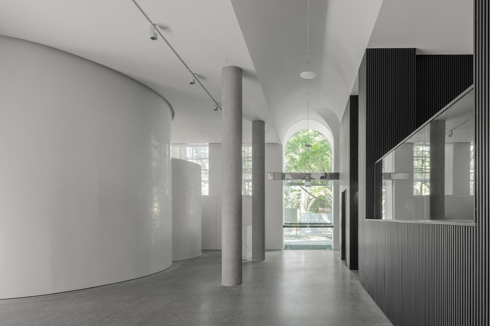 Interior corridor view through concrete columns toward arched window opening with greenery beyond