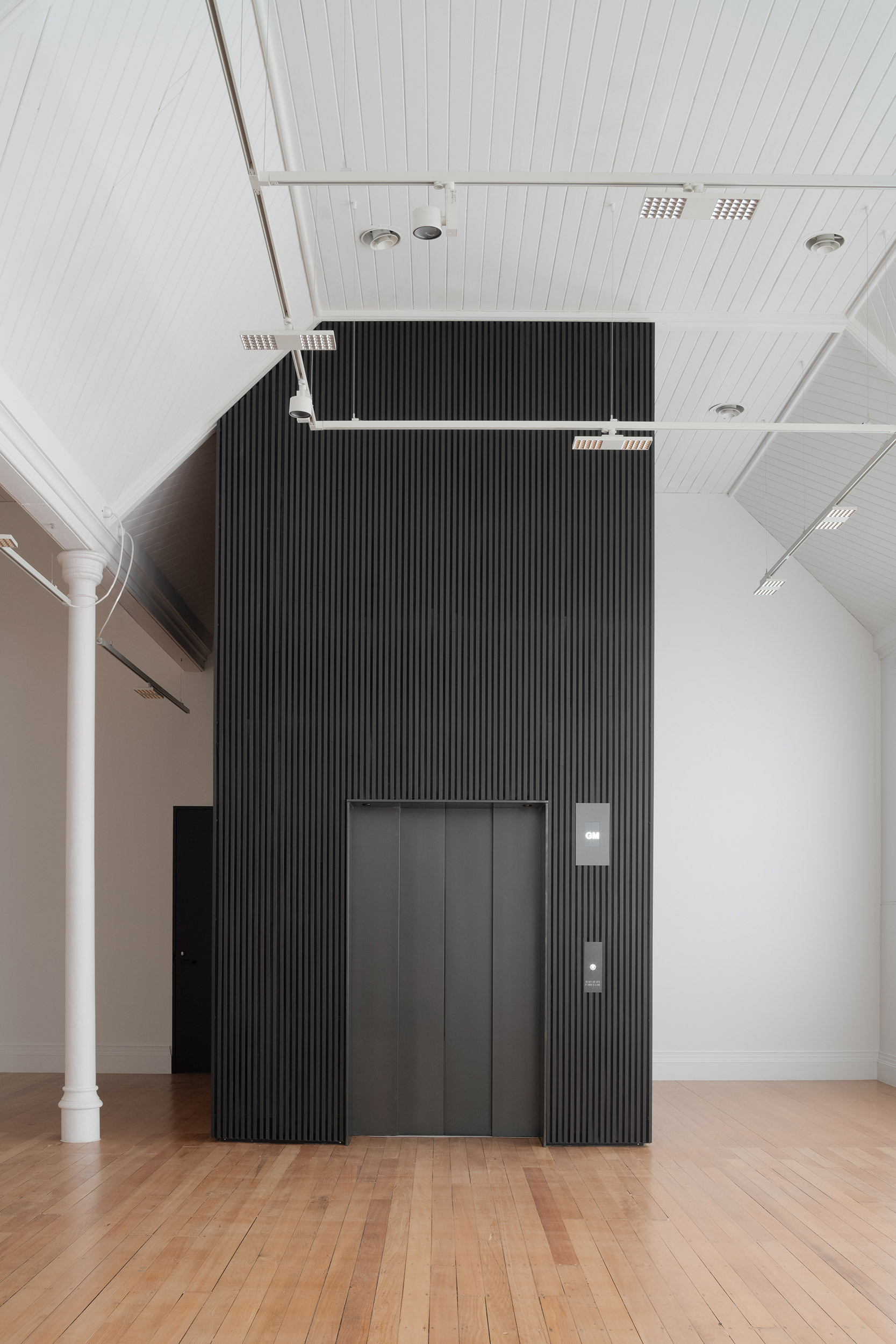 Black ribbed elevator core set against white paneled ceiling and timber floor in gallery hall