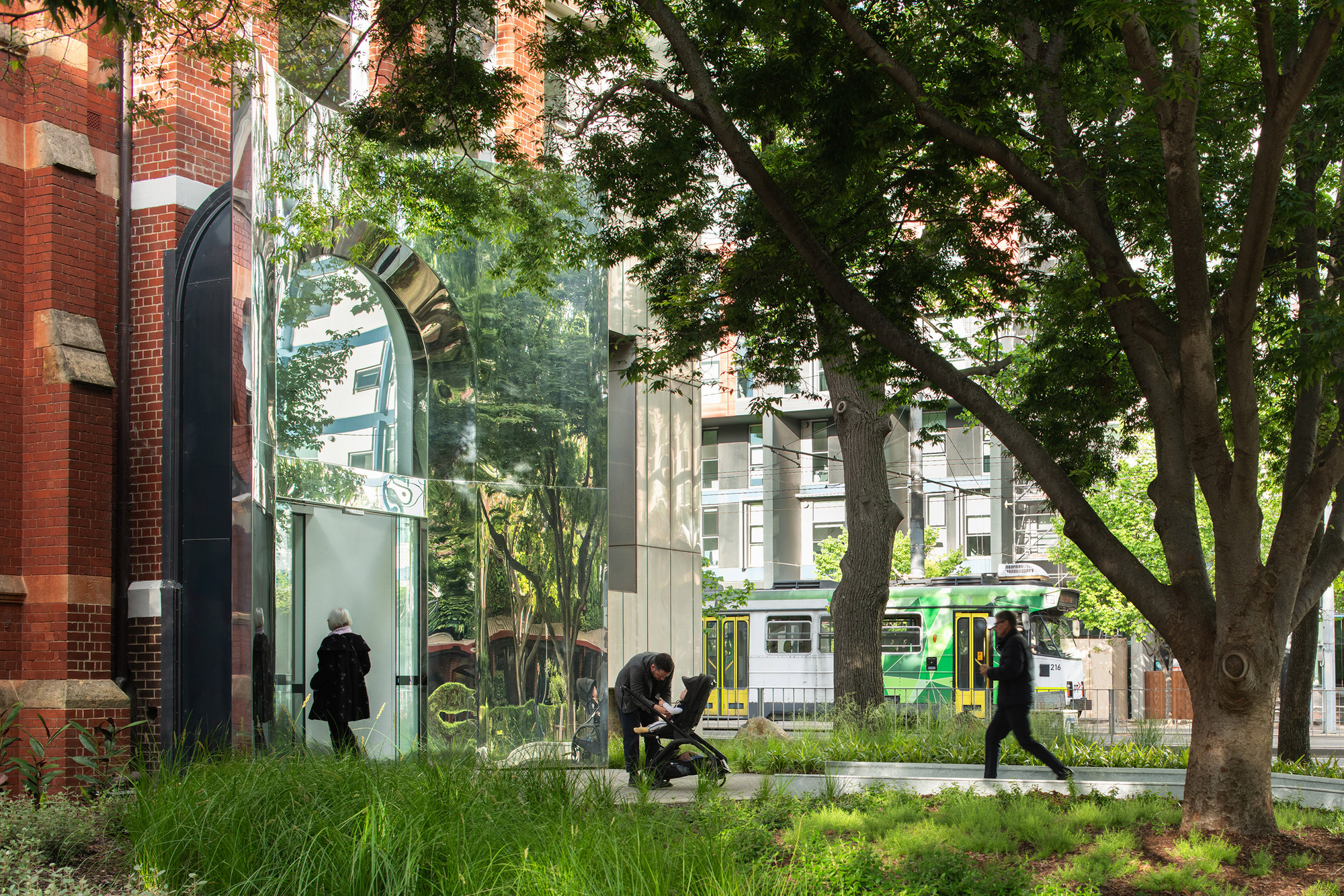 Street view of glass pavilion entrance among mature trees with pedestrians and passing tram