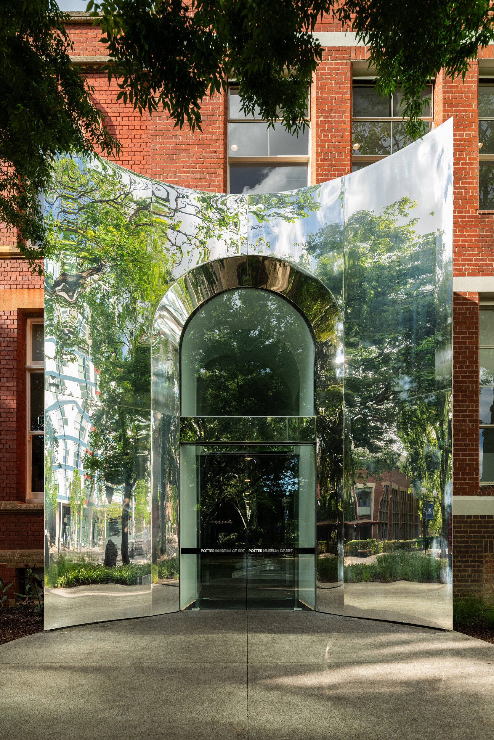 Curved glass entry volume with polished metal arch reflecting surrounding trees and brick walls in daylight