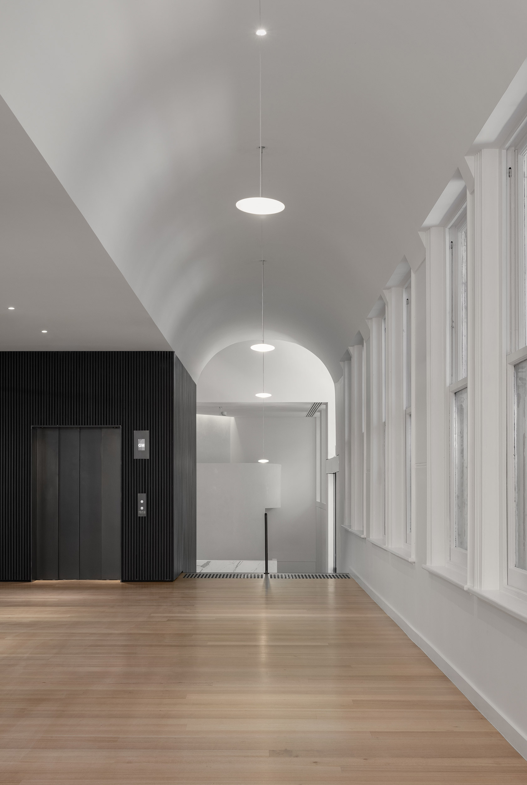 Corridor with vaulted ceiling, pendant lighting, timber floor and vertical black cladding around elevator enclosure