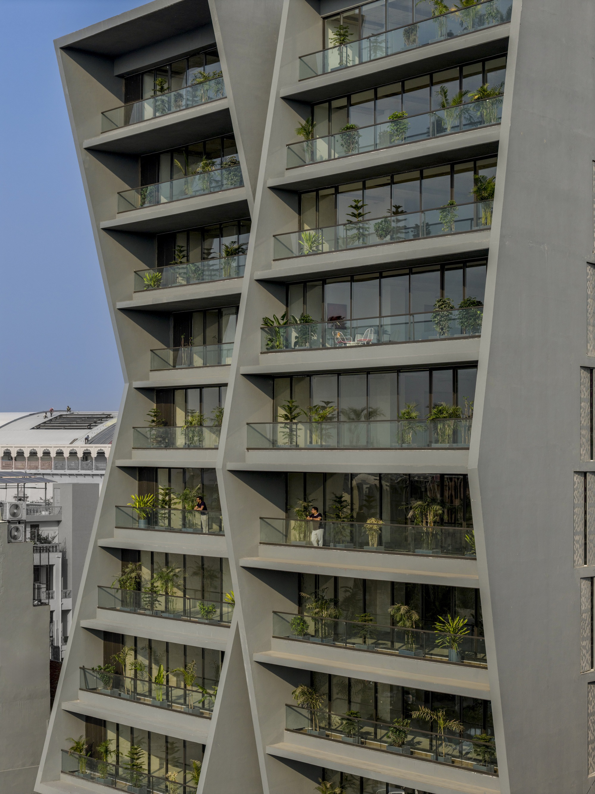 Diagonal facade with staggered balconies planted with greenery behind glass balustrades under clear sky