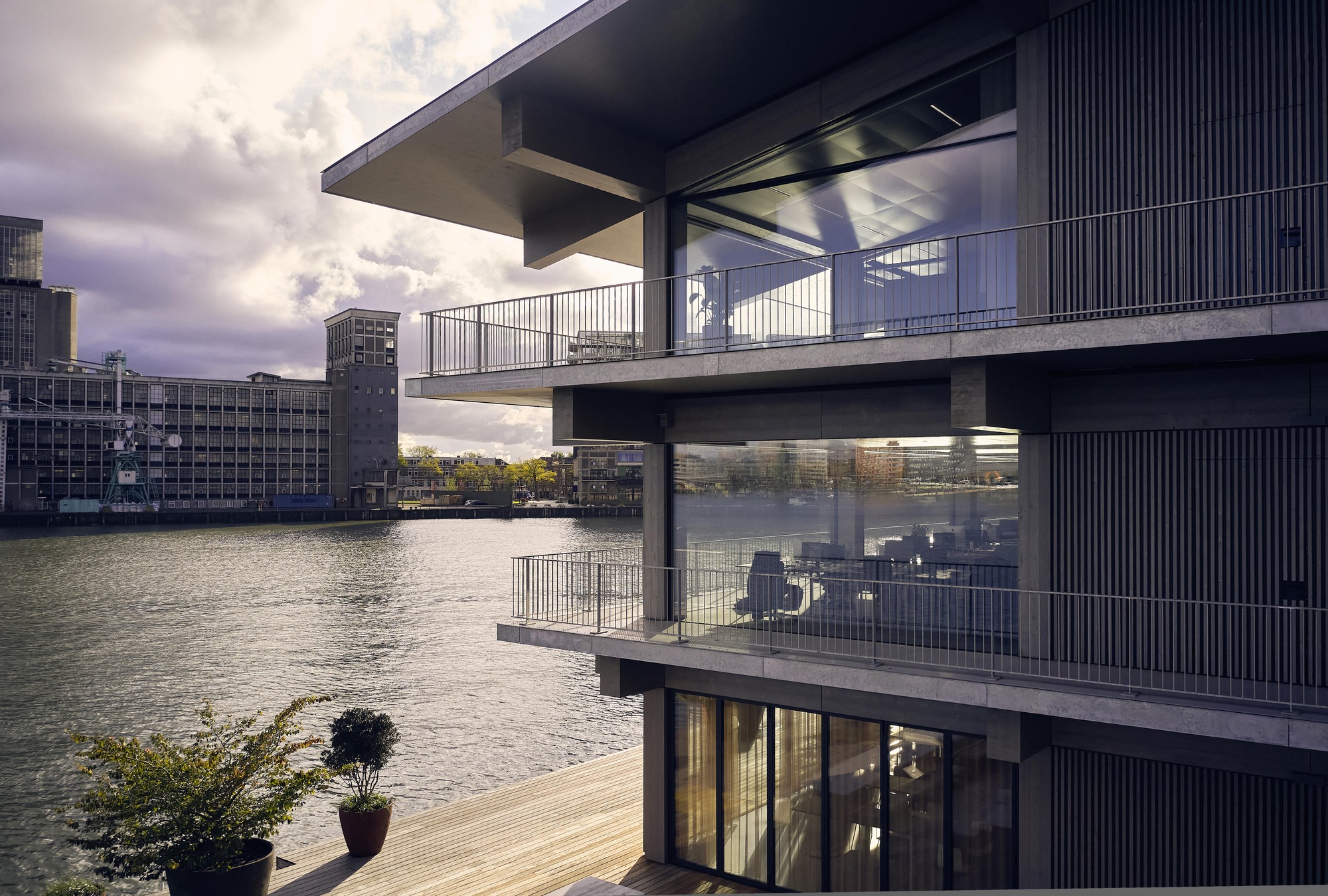 Waterfront facade with stacked concrete balconies, vertical metal screening and full-height glazing under stormy skies