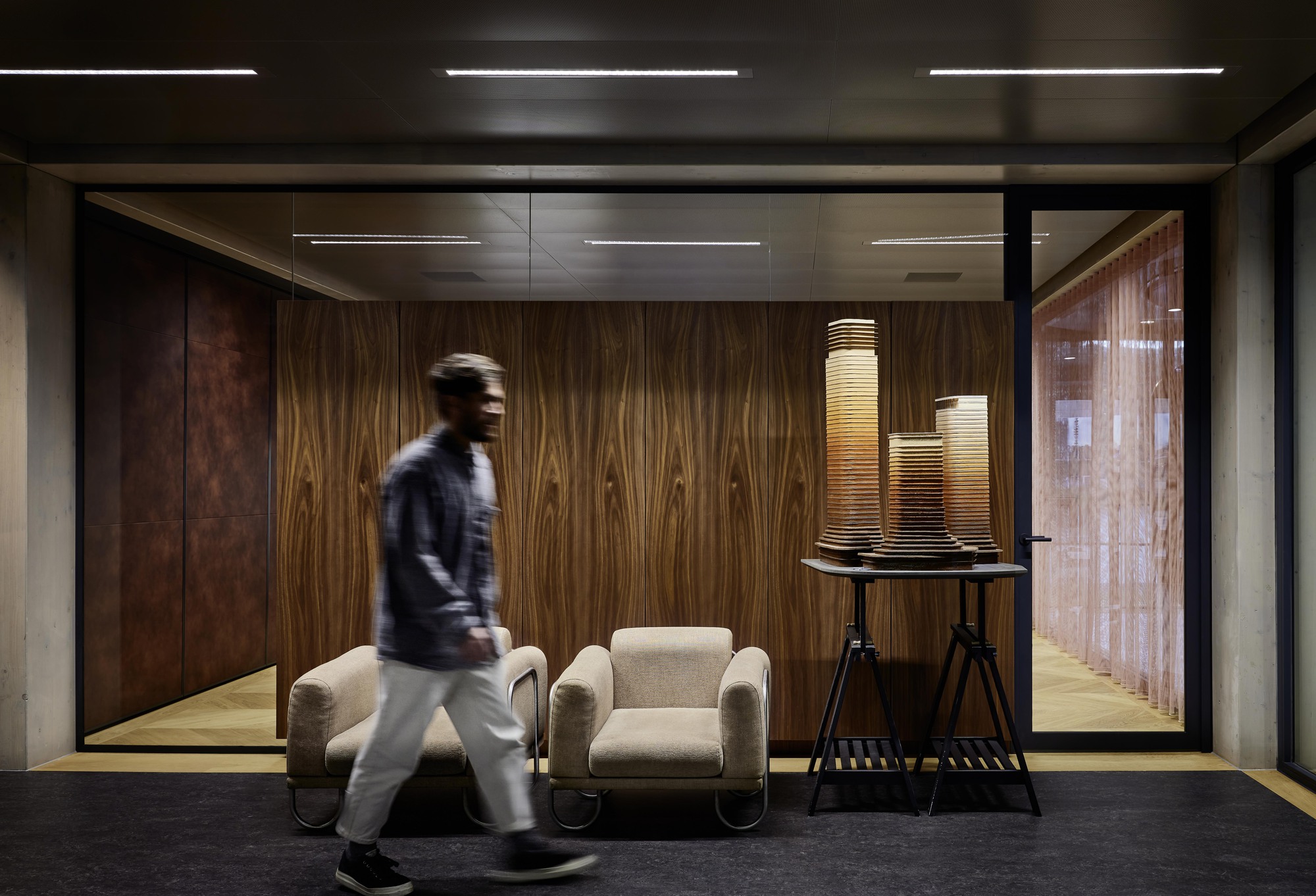 Wood-paneled corridor with glass partition and blurred figure walking past upholstered lounge chairs