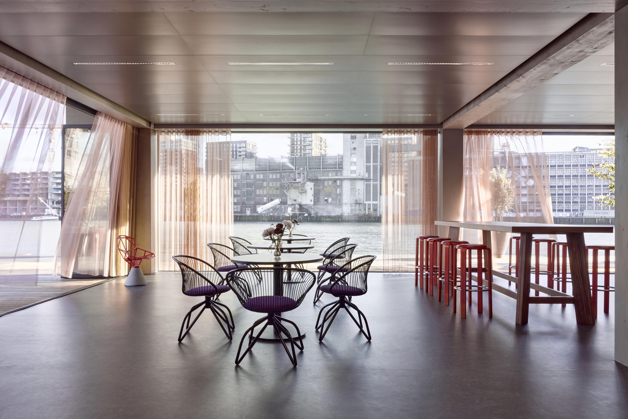 Dining area with metal frame chairs and floor-to-ceiling glazing overlooking a waterfront cityscape