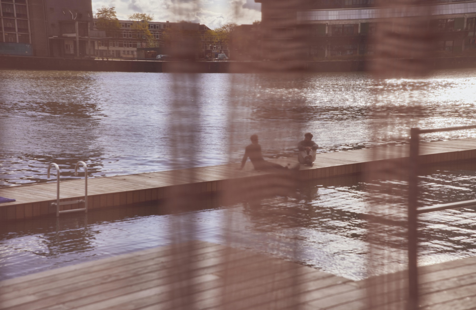 View through sheer curtains to timber swimming dock where two people sit in warm afternoon light