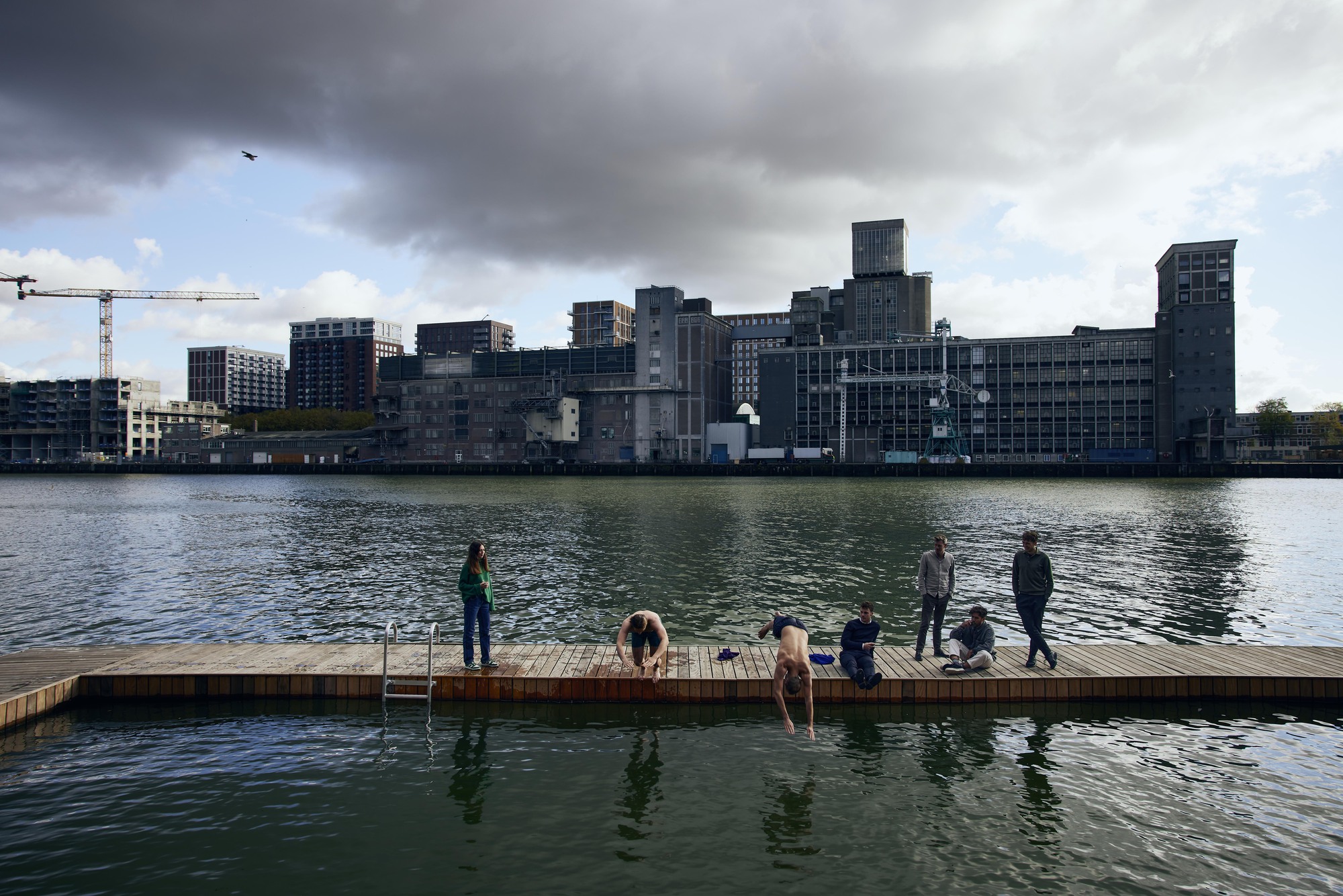 Timber swimming dock with people and a dog facing the converted industrial waterfront under dramatic clouds