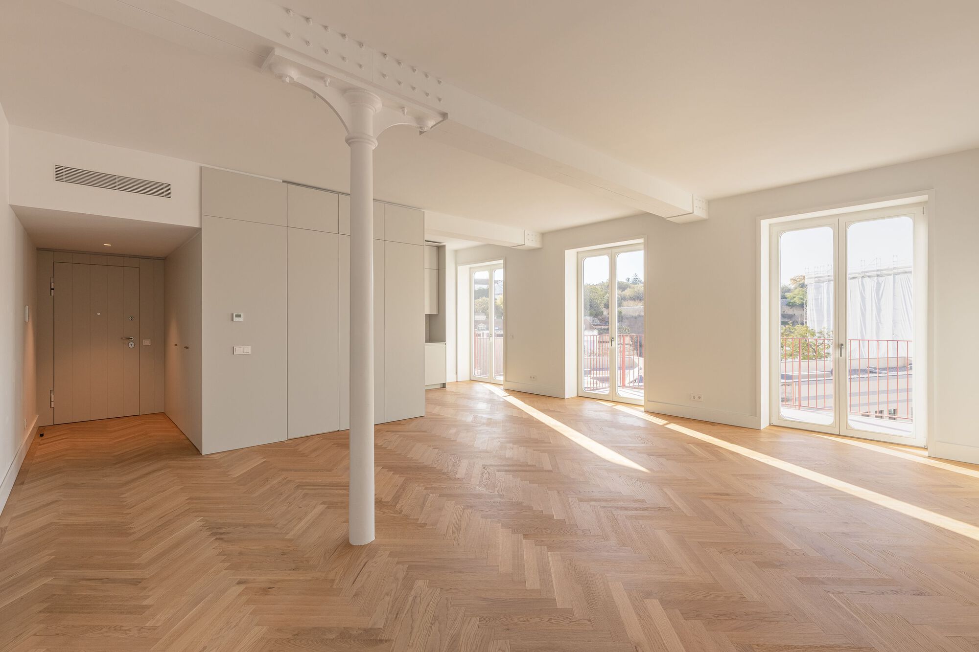 Open living space with herringbone oak flooring and a slender white column beneath sunlight