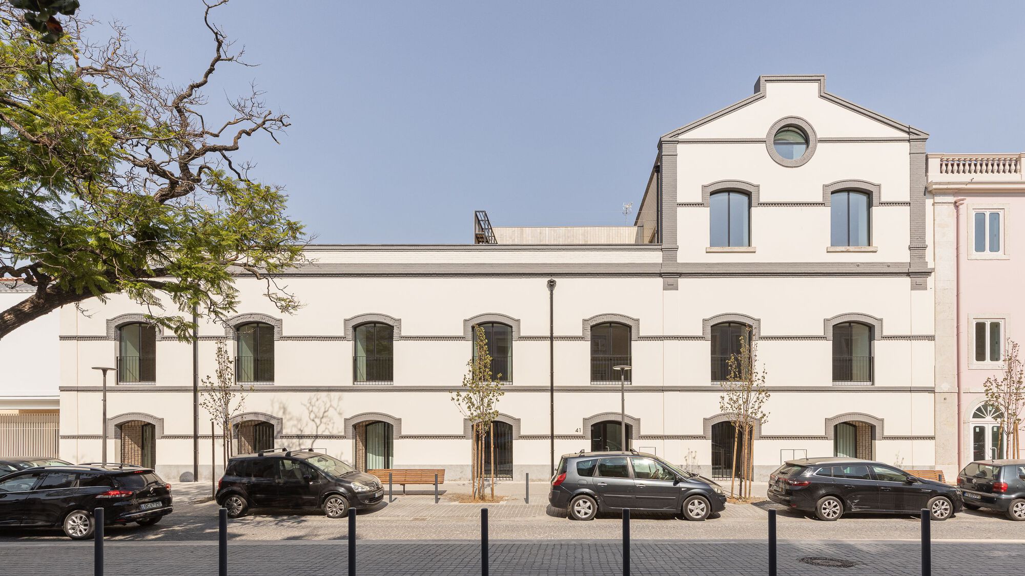 White rendered facade with arched windows and horizontal banding facing a public plaza with a mature tree