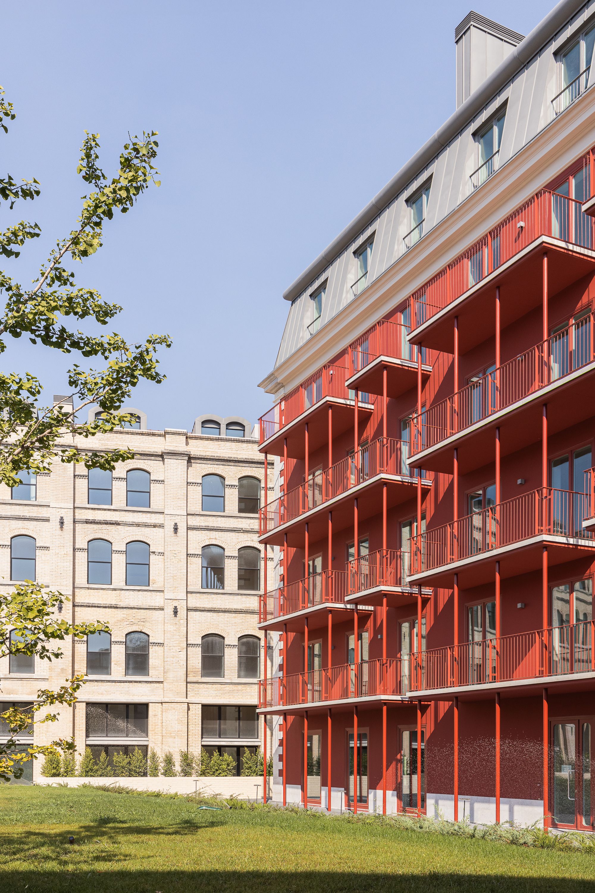 Red metal balconies stacked on a new block facing a historic arched brick building across a lawn