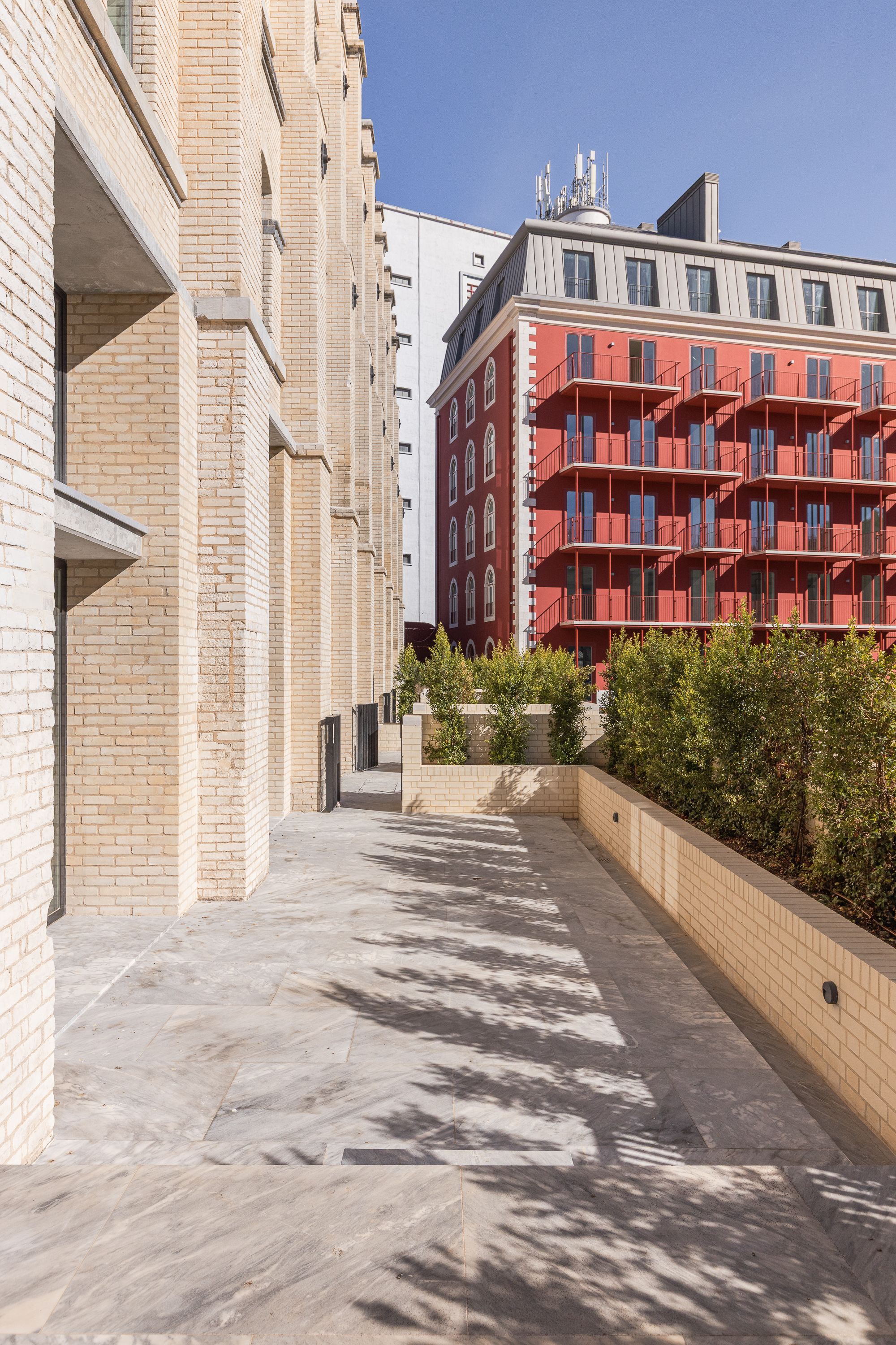 Paved pedestrian pathway between cream brick wall and planted beds with red residential block in background
