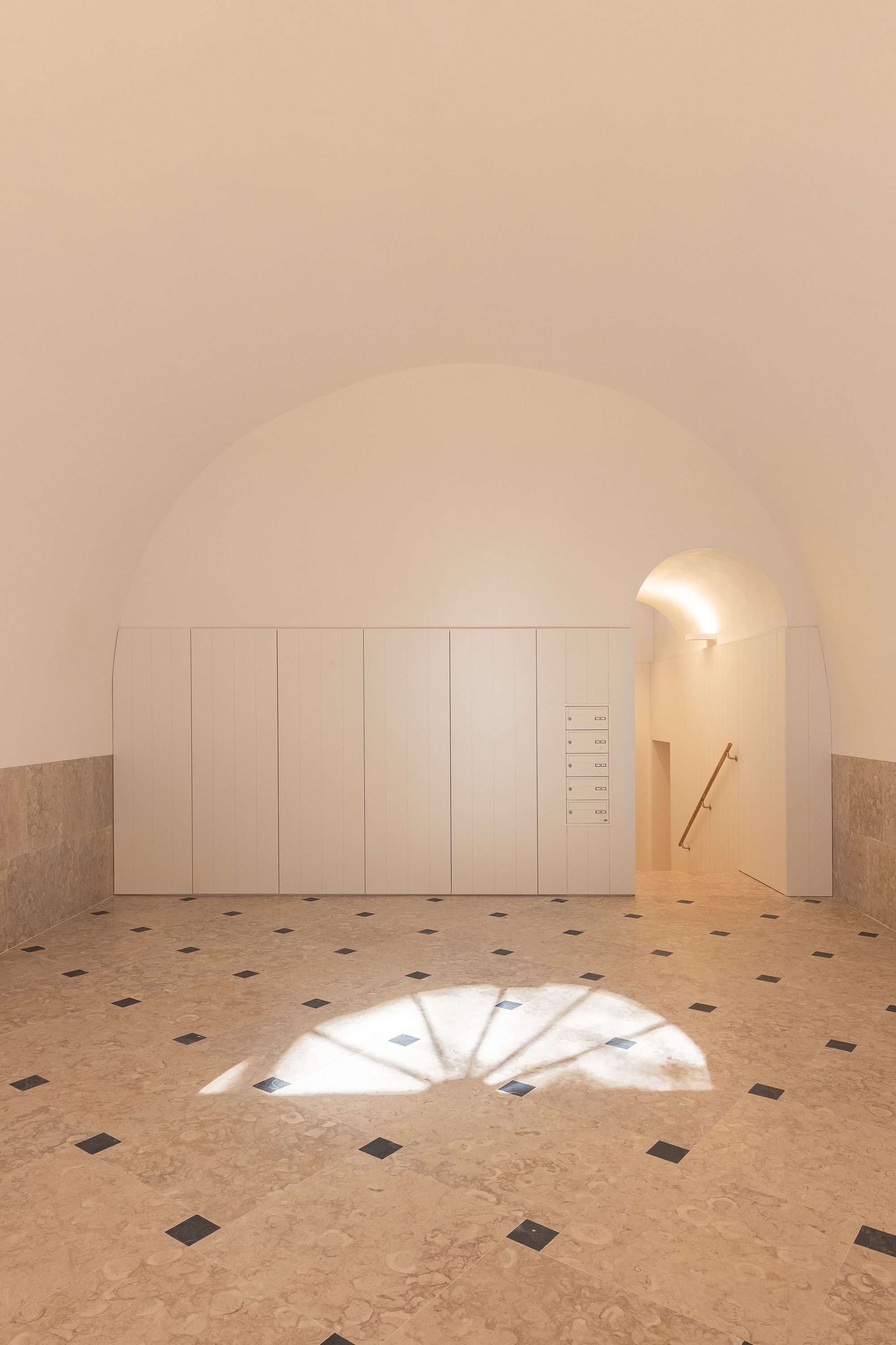 Vaulted entry space with white paneled wall and patterned terrazzo floor casting arched window shadows