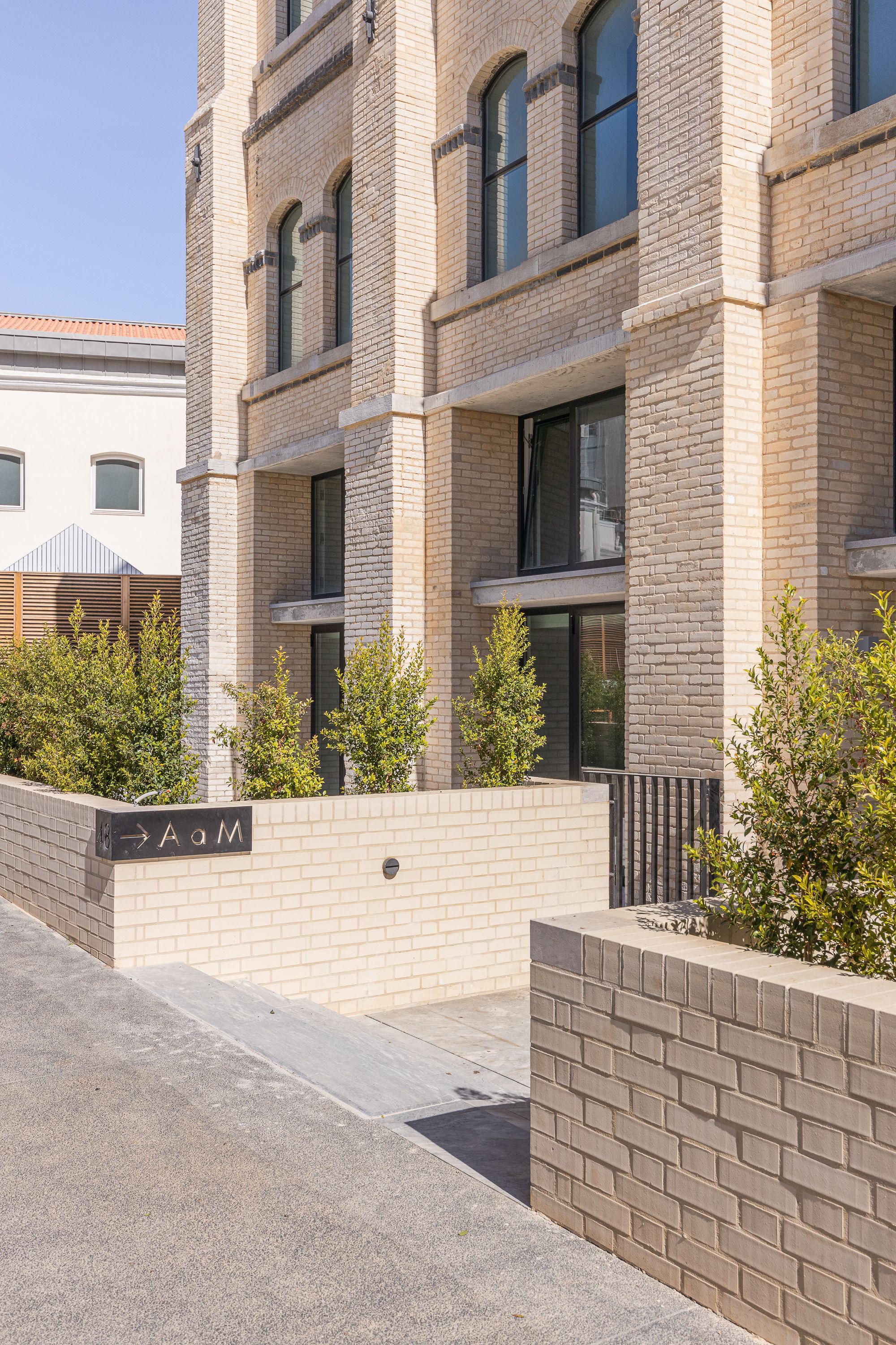 Buff brick facade with arched windows above a landscaped entry wall with young trees in afternoon light