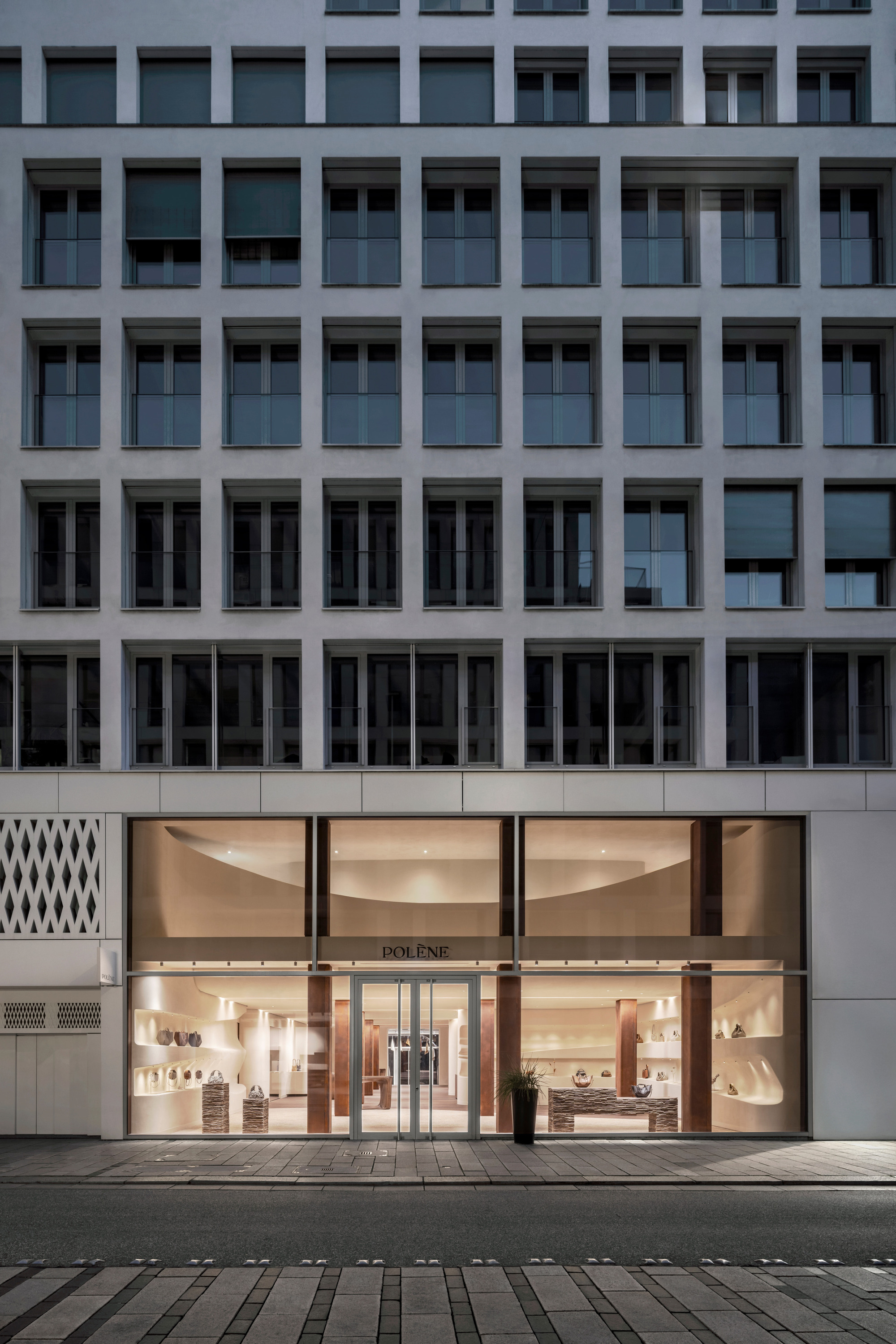 Street-level storefront with curved glass facade illuminated at dusk beneath a grey office tower