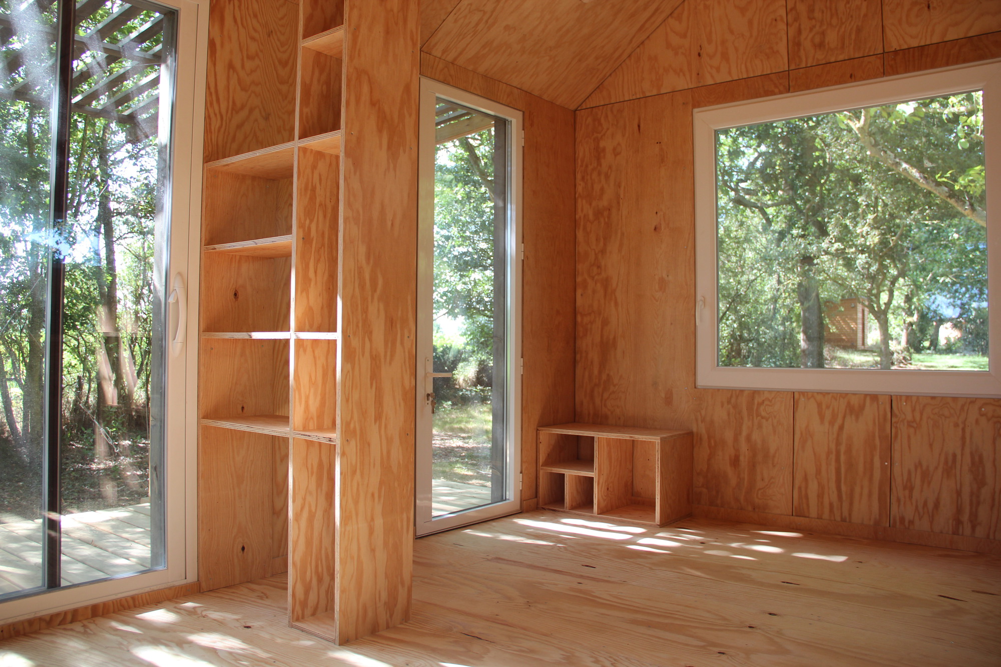 Interior plywood-lined room with built-in shelving and glazed doors opening to the deck in afternoon light