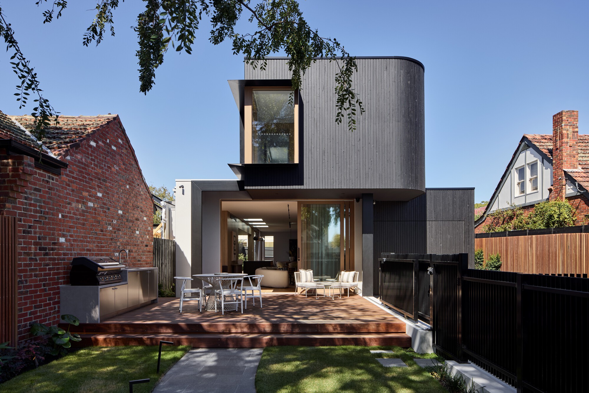Rear garden view showing timber deck with outdoor dining and cylindrical upper volume clad in grey ribbed panels