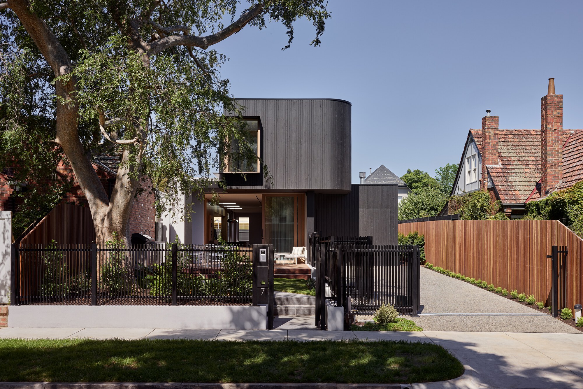 Street view of the black corrugated metal facade with vertical slat fencing and a mature tree overhead