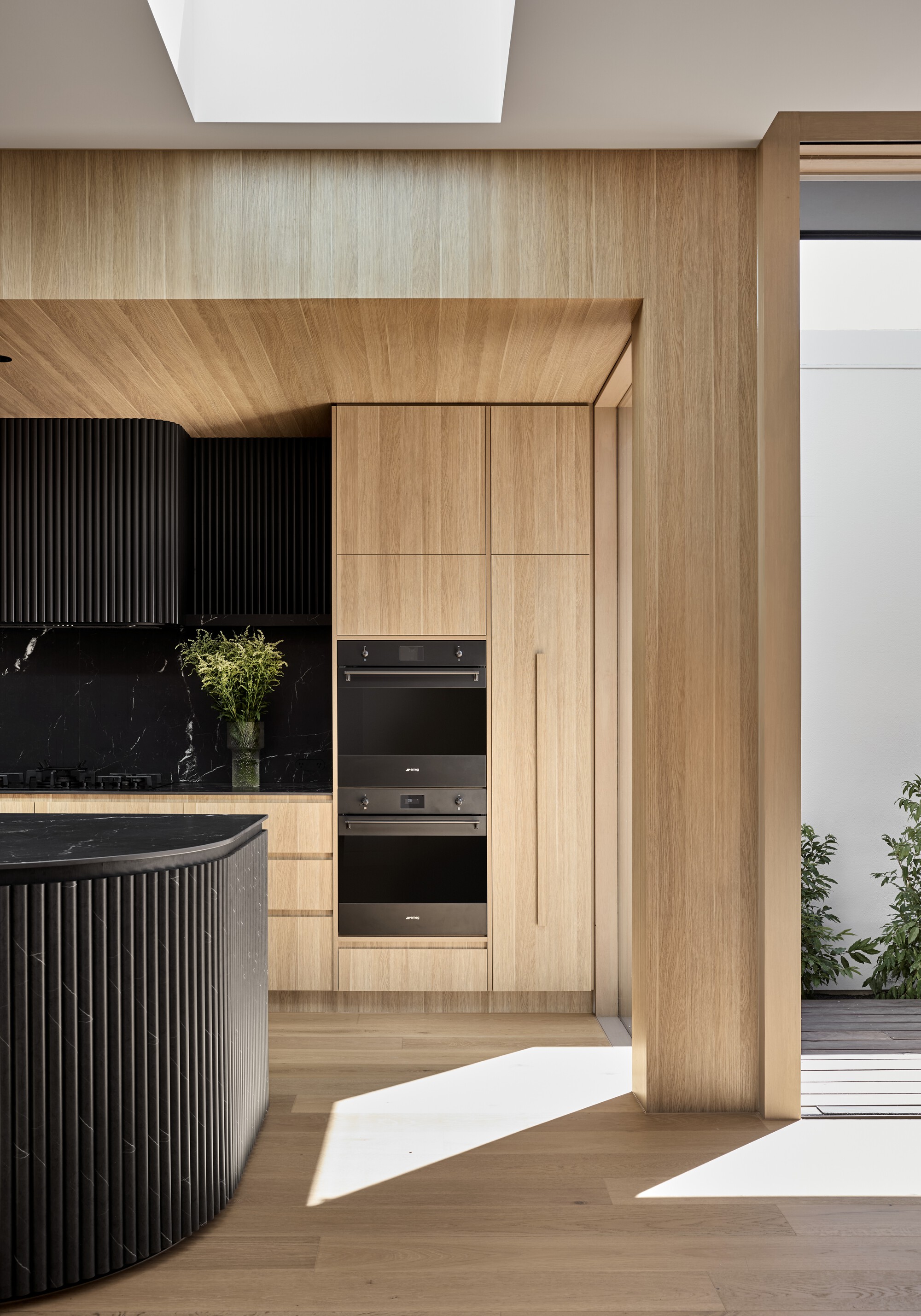 Kitchen view toward built-in ovens framed by timber portal with sunlight streaming across the floor