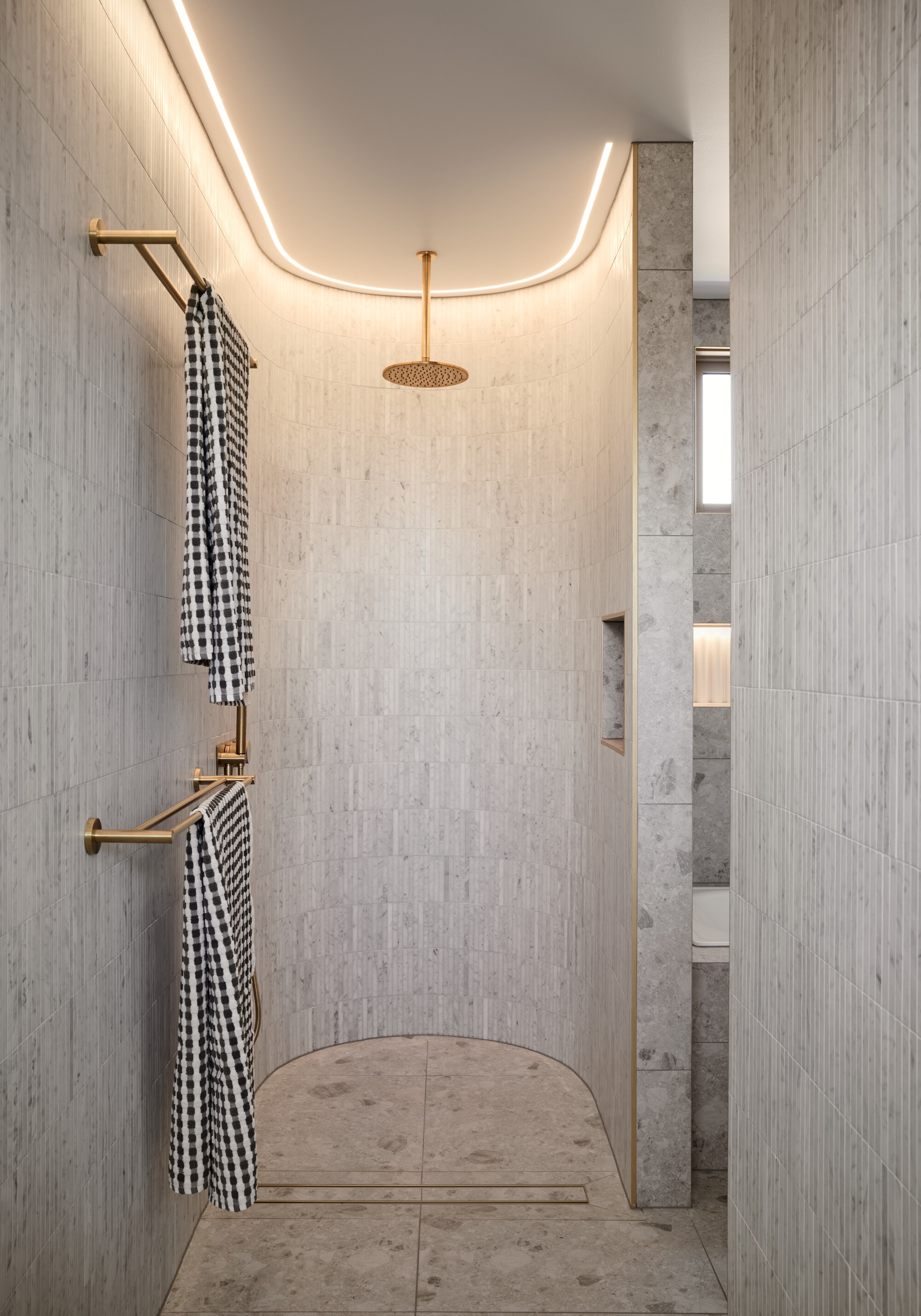 Curved shower niche with integrated ceiling lighting and brass rainfall head above patterned floor tile