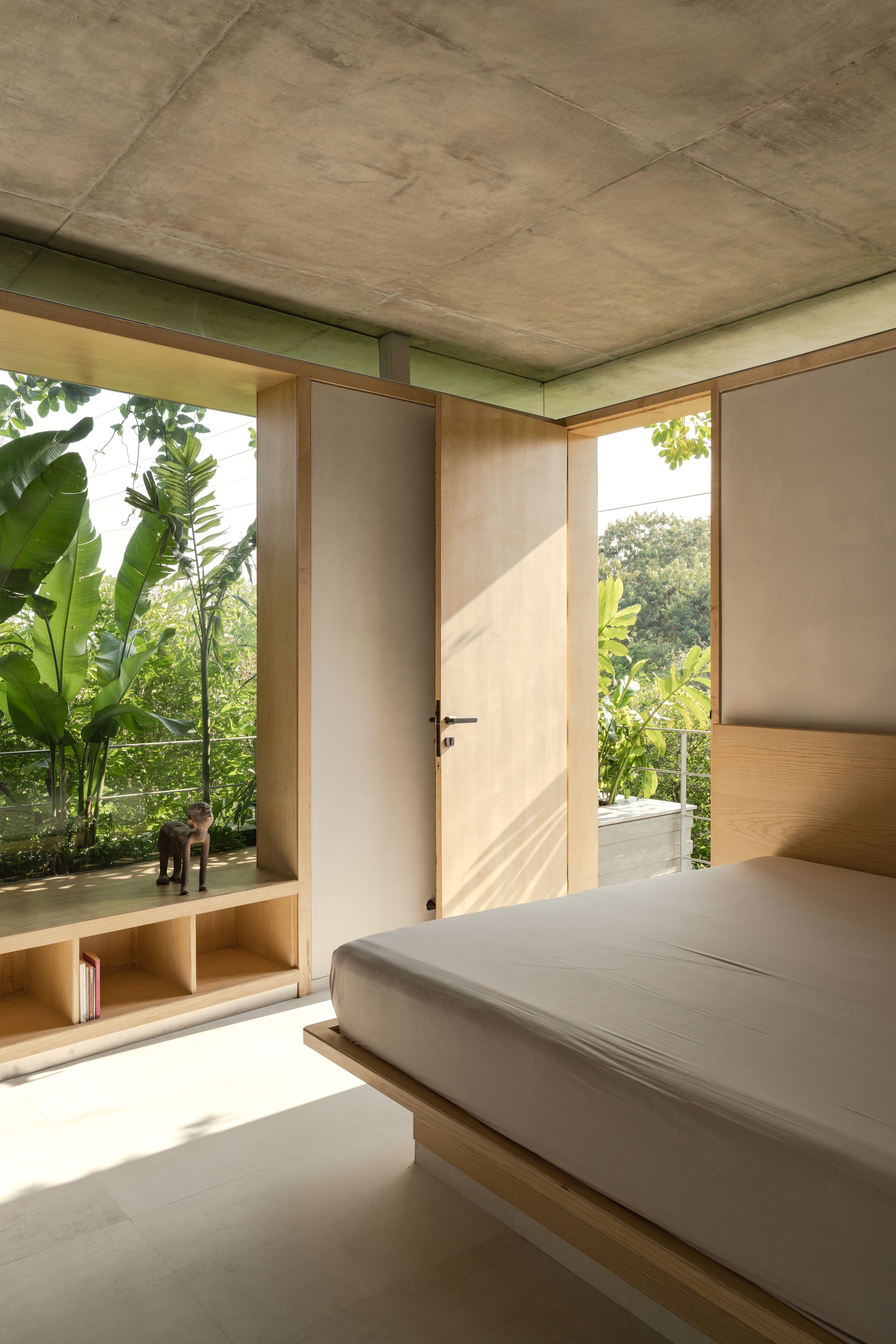 Bedroom with exposed concrete ceiling, built-in timber bed platform, and full-height glazing overlooking tropical vegetation