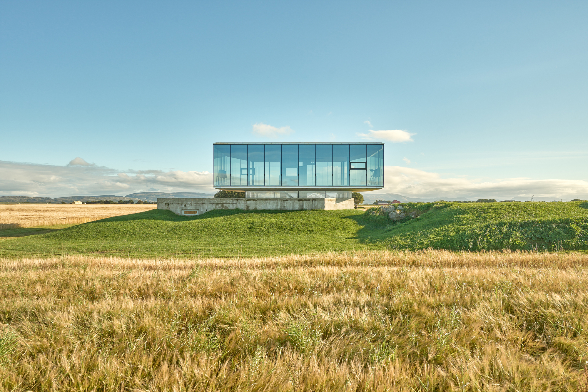 Glass volume cantilevered above a grass-covered berm in a golden wheat field under afternoon light