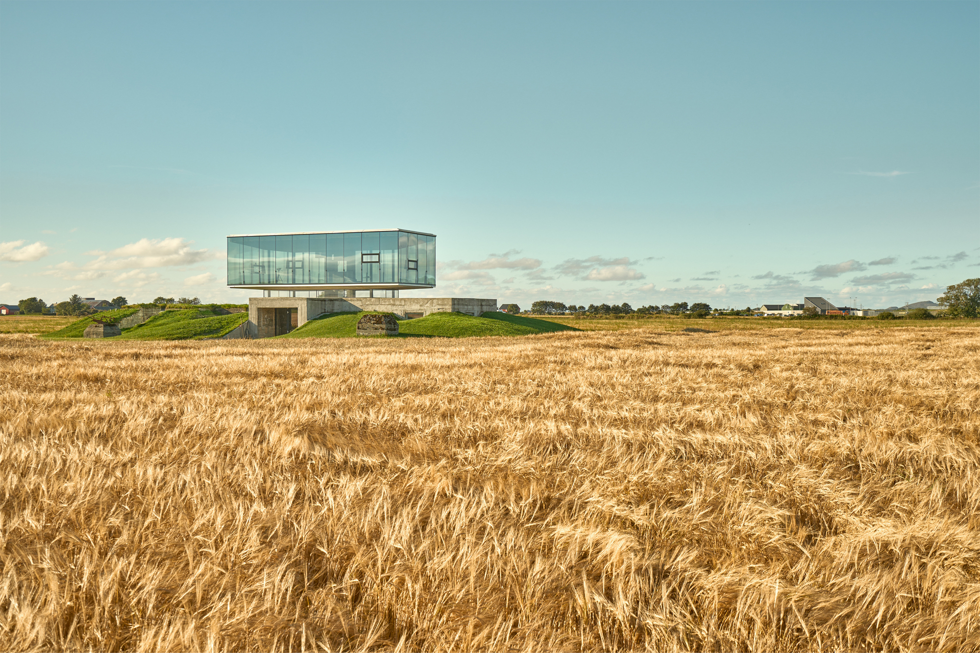 Elevated glass pavilion on concrete supports rising from a harvested wheat field
