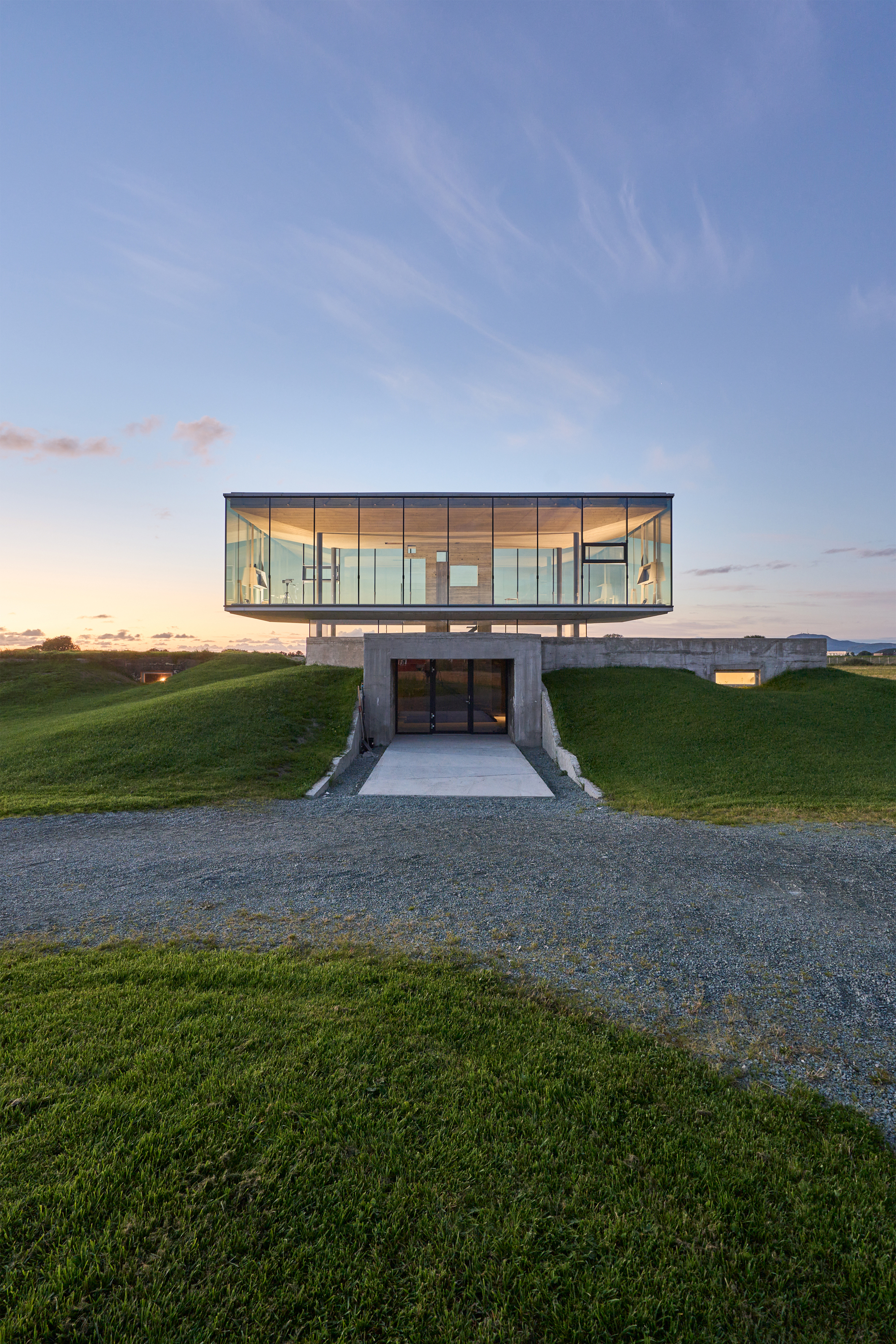Frontal view of the glass volume cantilevered above a recessed concrete entrance at dusk