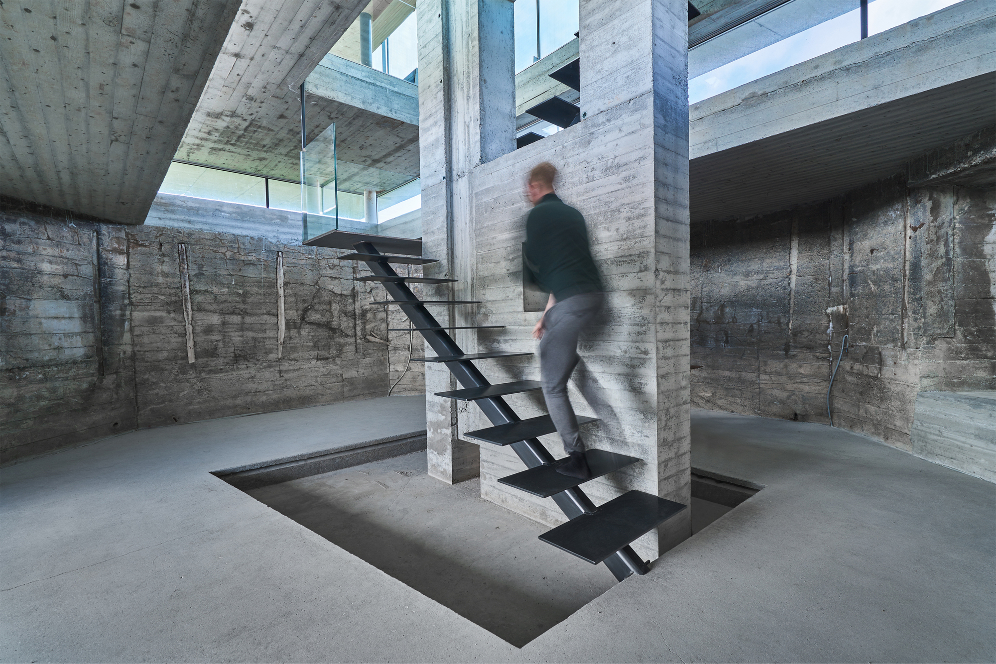 Board-formed concrete interior with a black steel staircase wrapping a column as a figure ascends