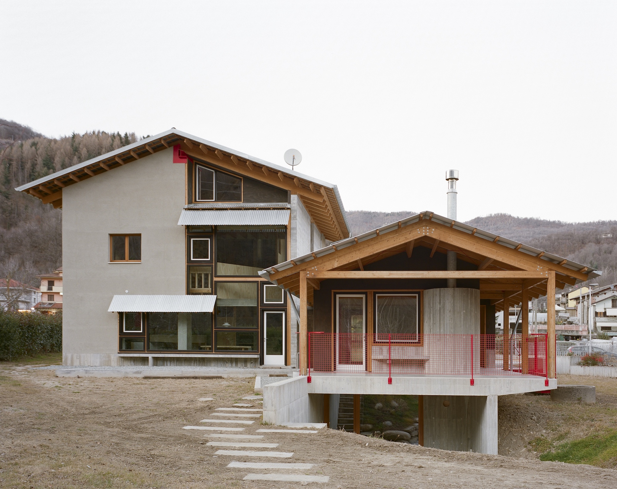 Two-volume composition with exposed timber eaves and red mesh railings set against a hillside village