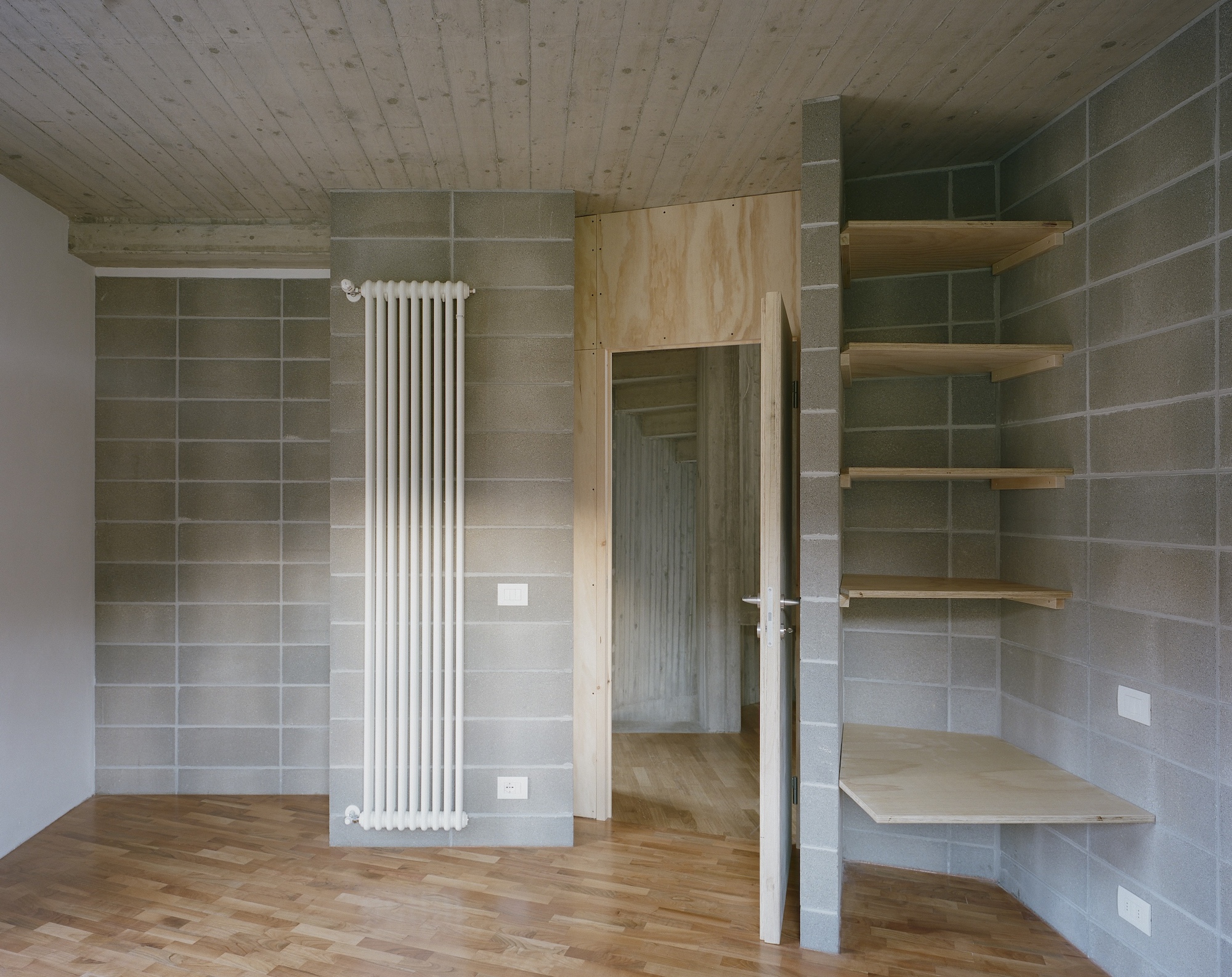 Corner view showing plywood door, radiator, and floating shelves against concrete block walls
