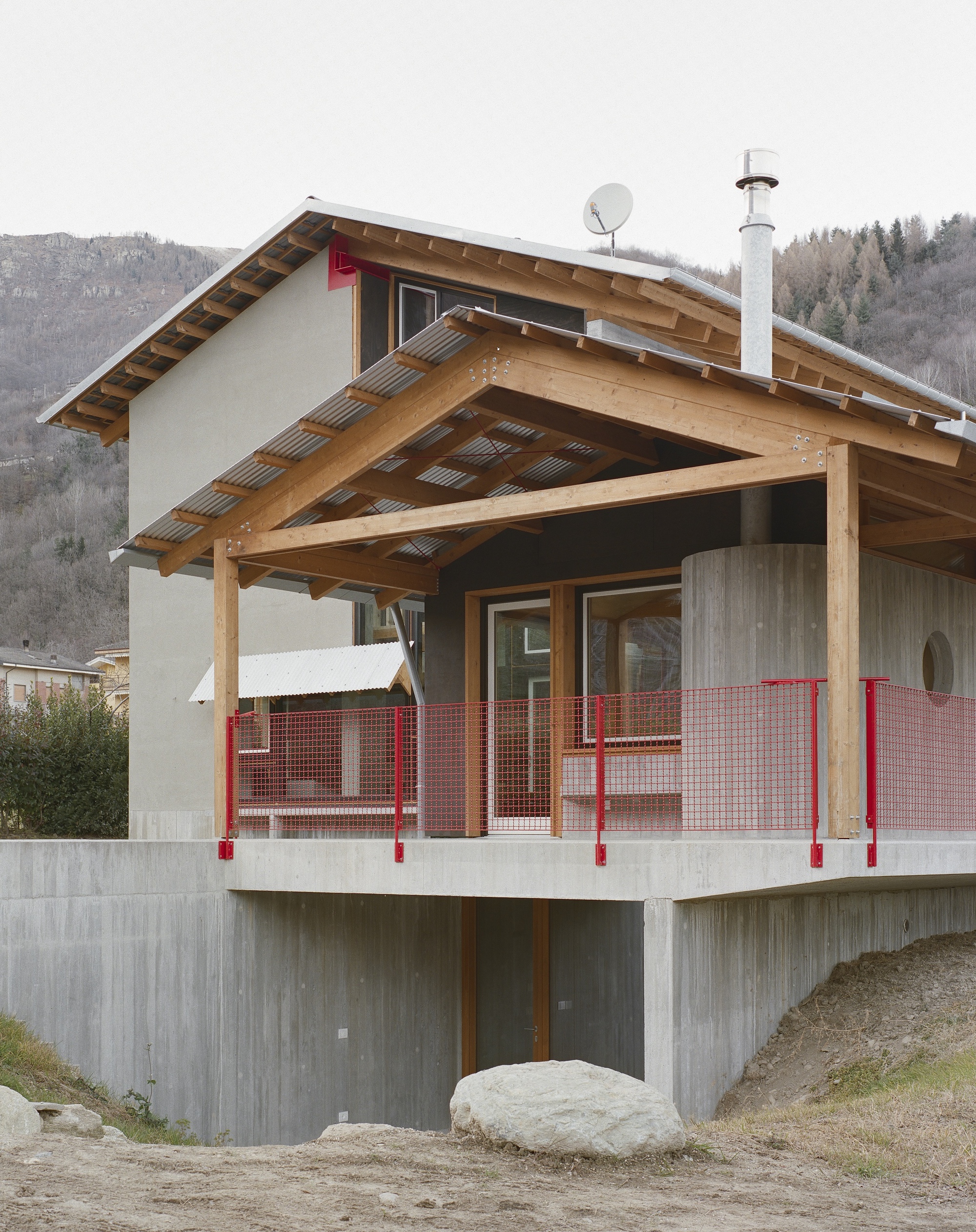 Timber-framed porch with exposed rafters and red mesh railing atop a concrete plinth base