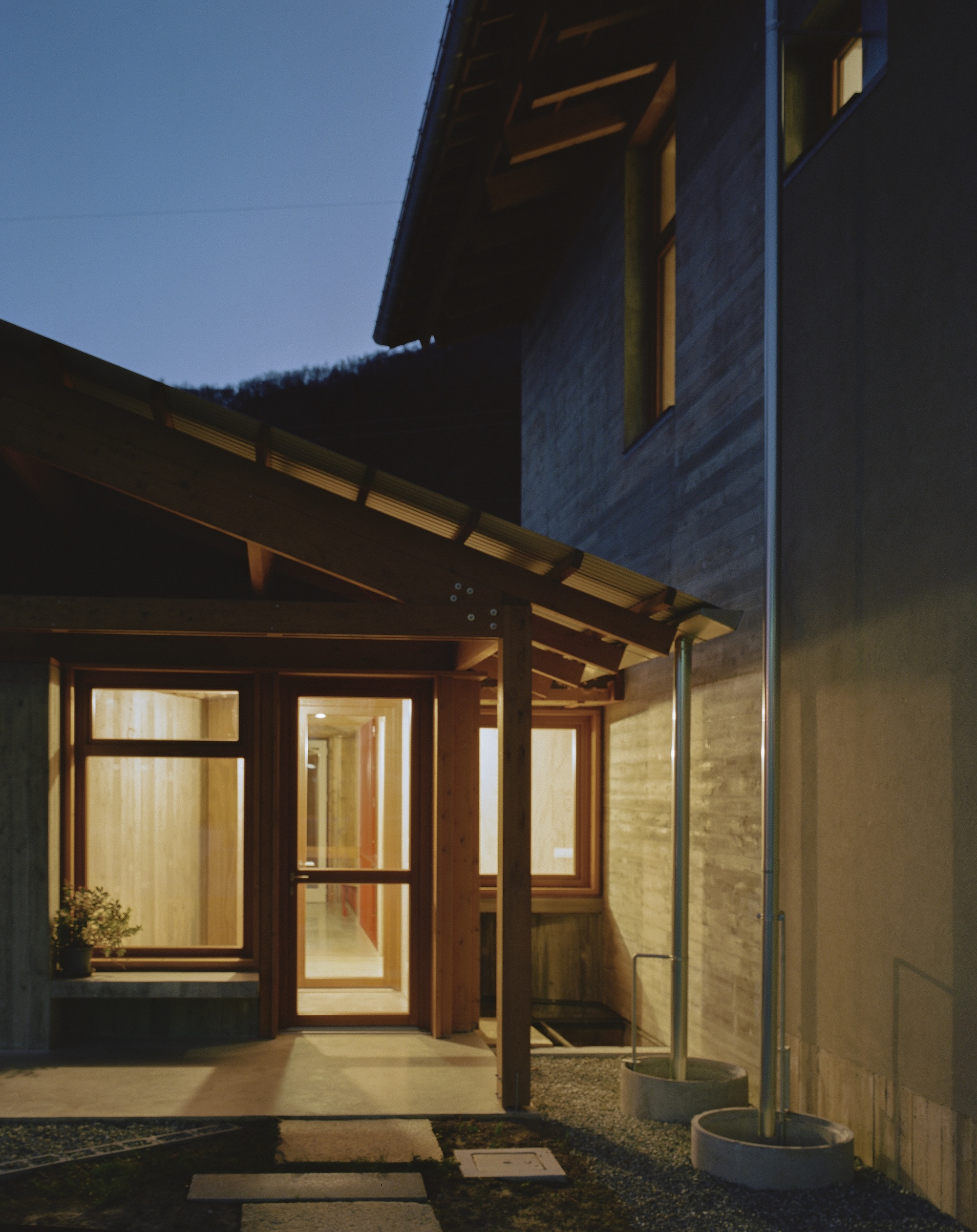 Illuminated timber entry pergola with exposed joists connecting concrete and plaster volumes at dusk