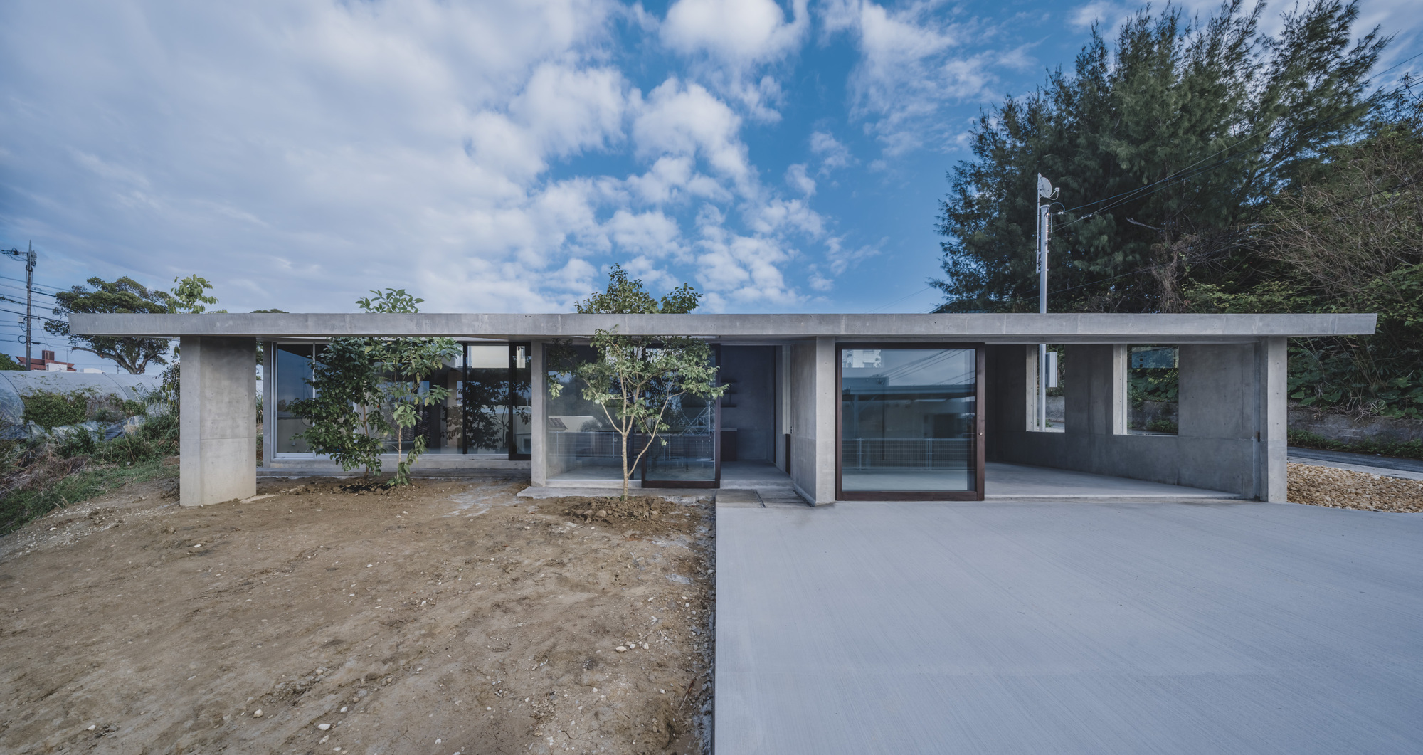 Front facade with concrete columns framing glazed openings and young planted trees under blue sky
