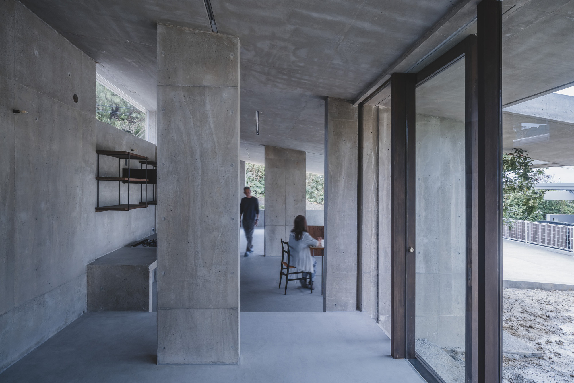 Two figures inhabit an open concrete interior with freestanding columns and a wall-mounted shelving unit