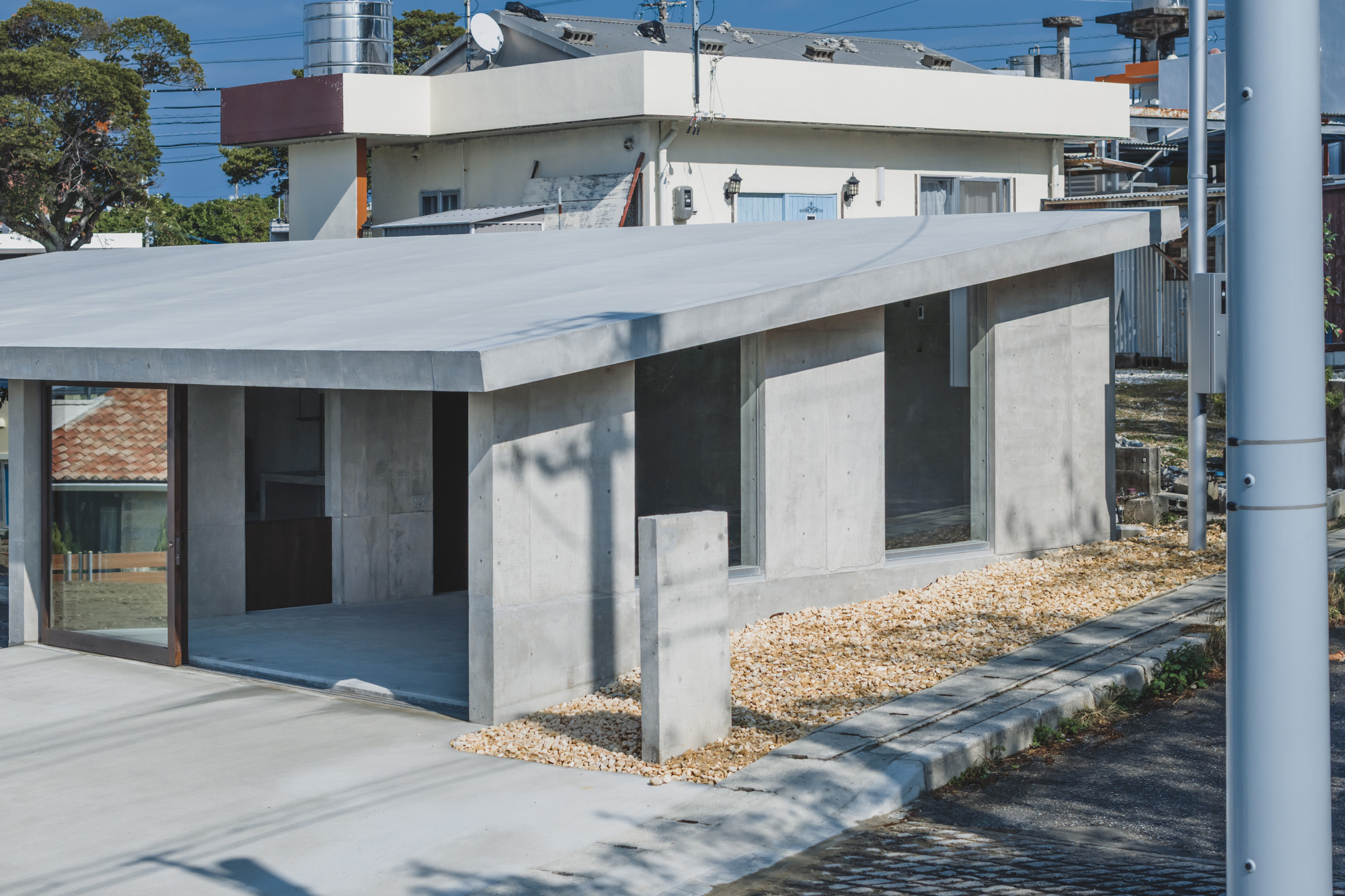 Concrete volume with flat overhang sheltering a recessed glazed opening above a gravel yard in daylight