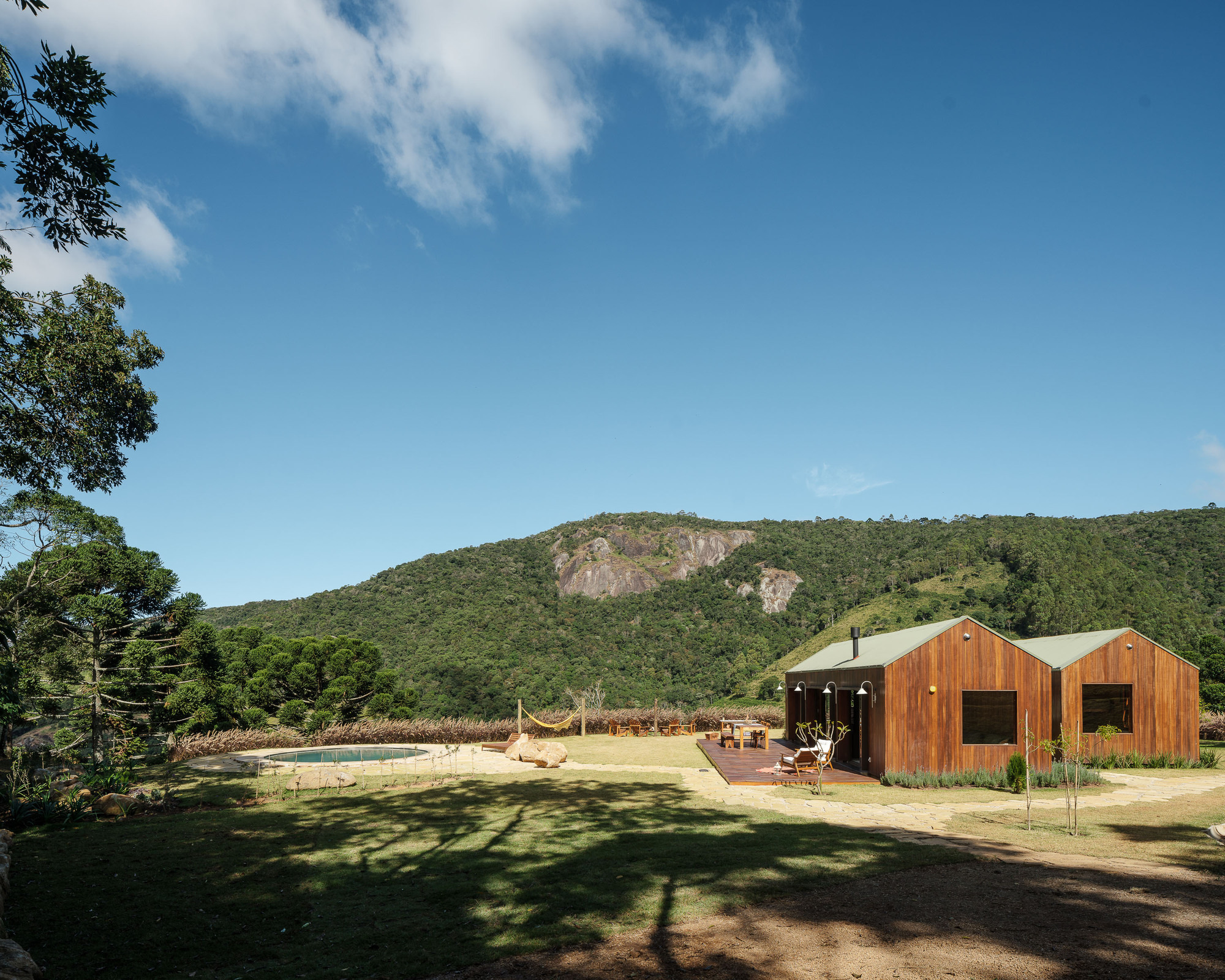 Two gabled timber pavilions with metal roofs anchoring a lawn clearing beneath forested hillside