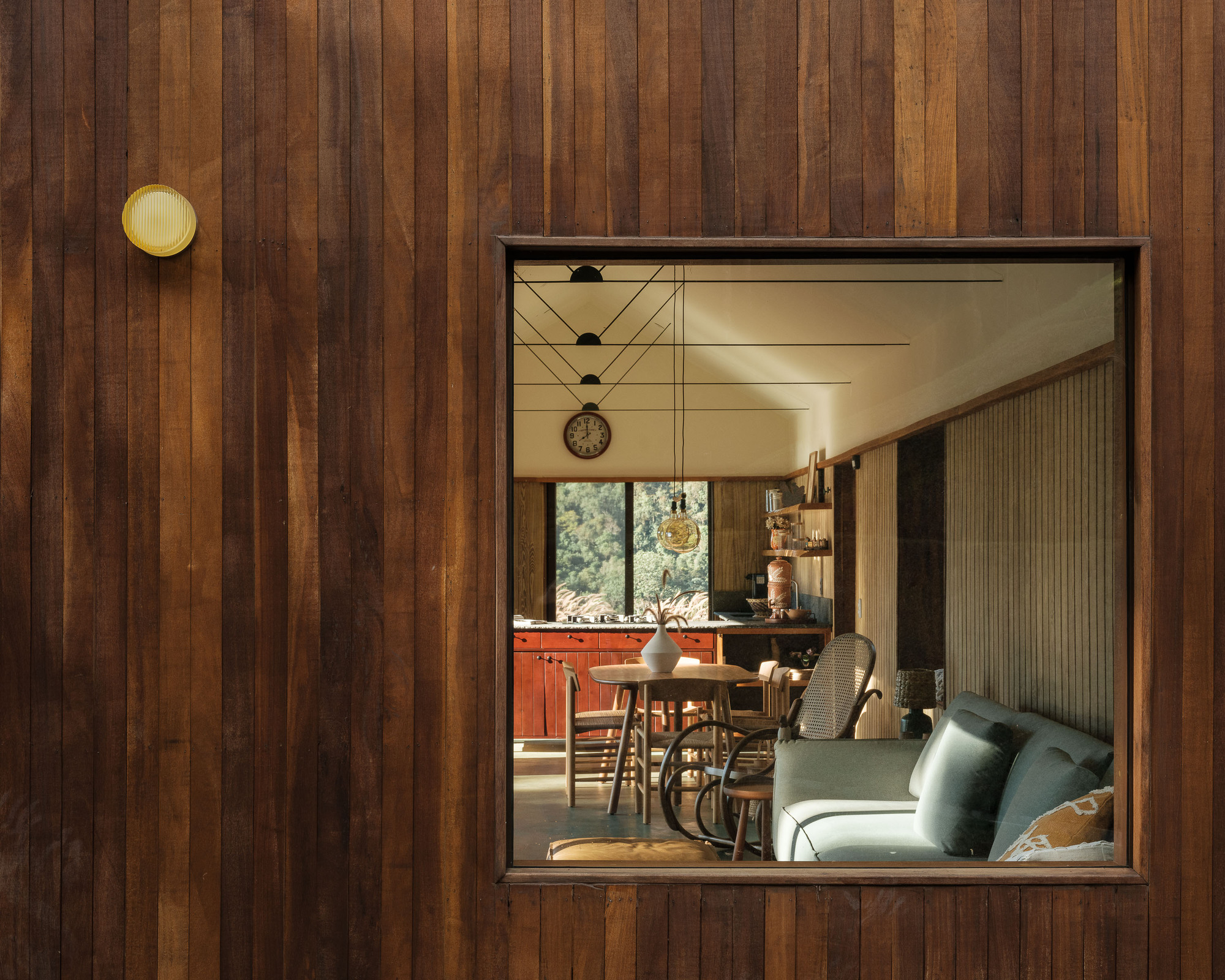 Square window framed in vertical timber cladding reveals the dining area with pendant lights and a sofa