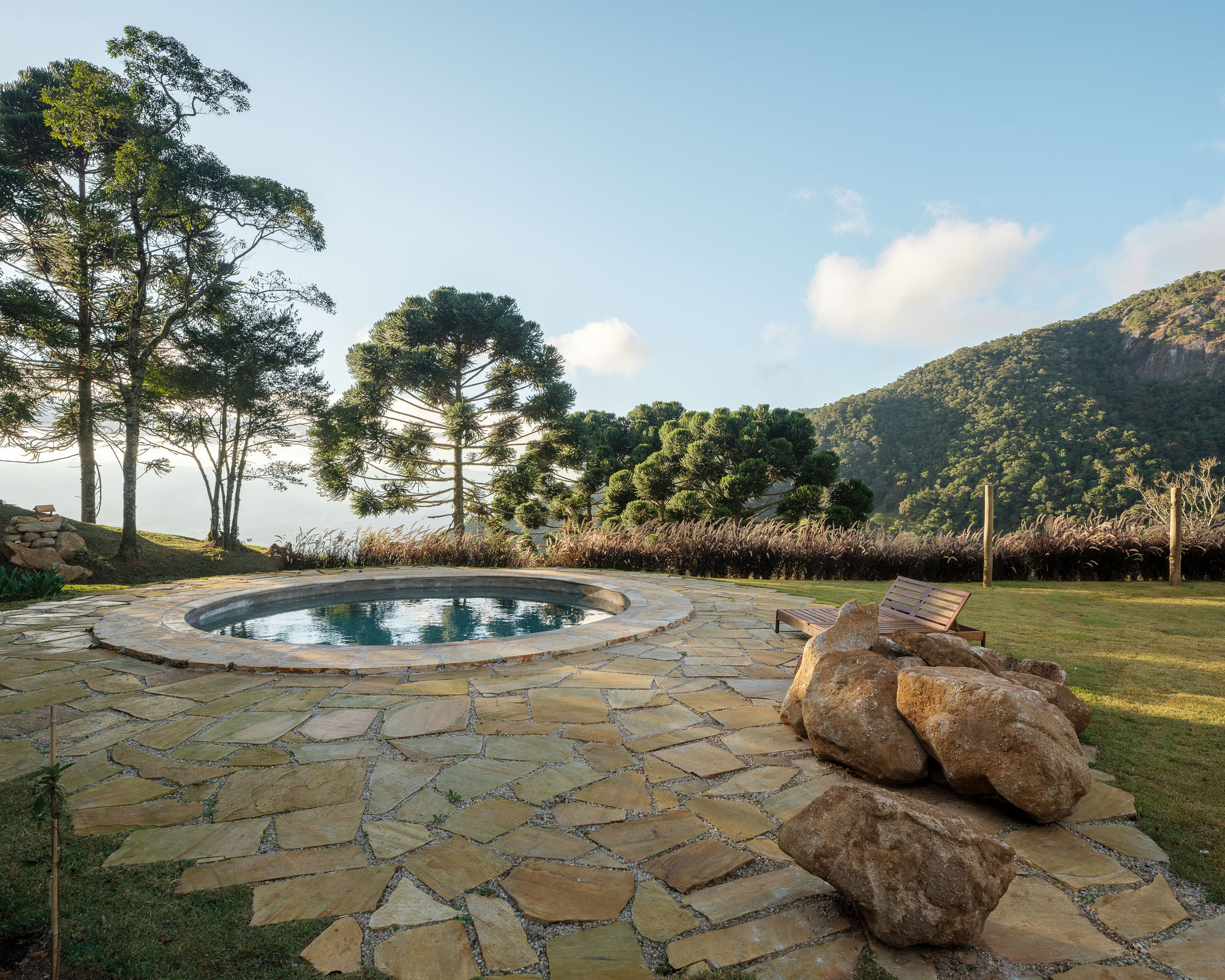 Circular pool surrounded by flagstone paving with boulders and grasses facing forested hills