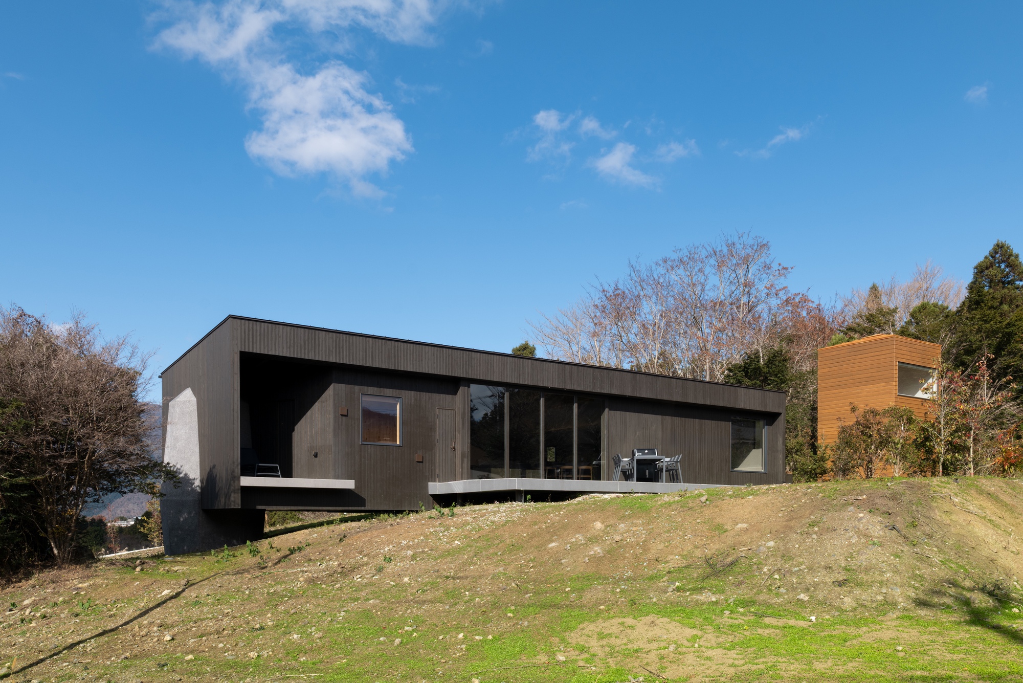 Dark timber-clad volume with cantilevered terrace perched on a grassy hillside slope under a blue sky