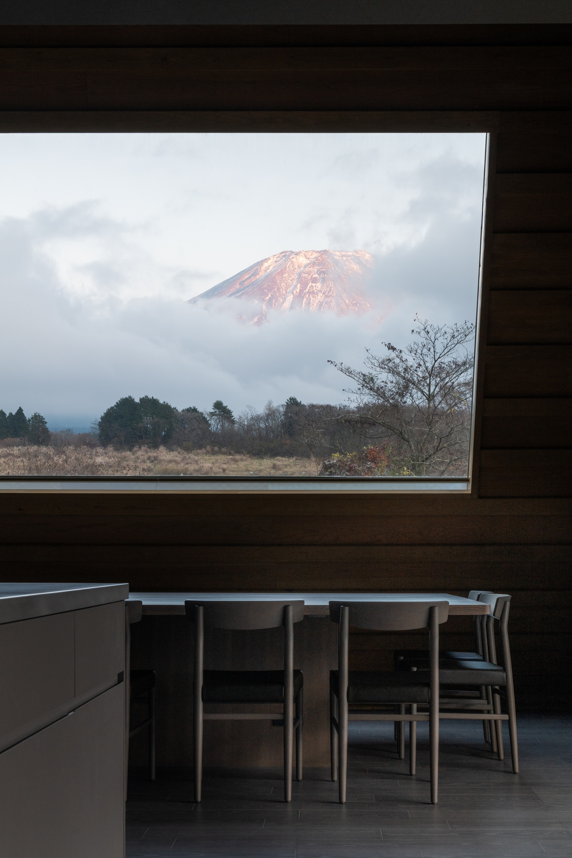 Horizontal window in timber-clad dining space framing a distant snowcapped mountain through clouds