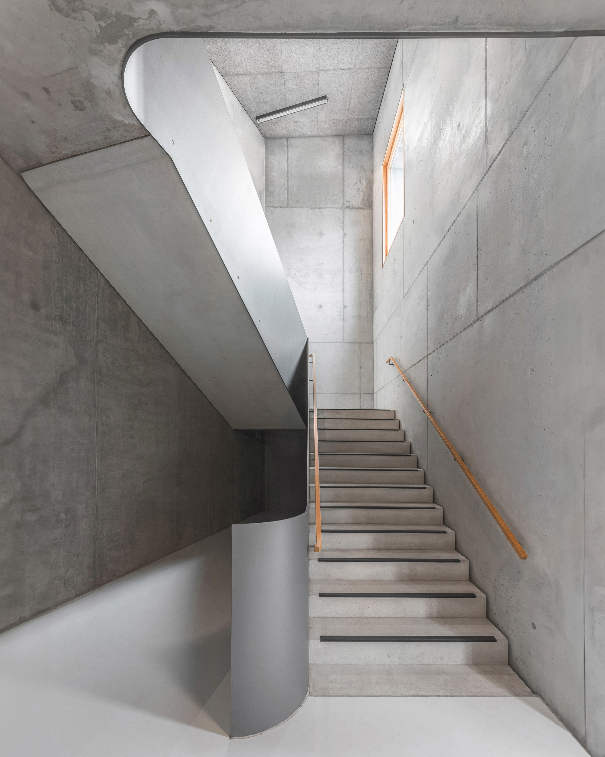 Concrete stairwell with curving white balustrade and timber handrail under natural skylight