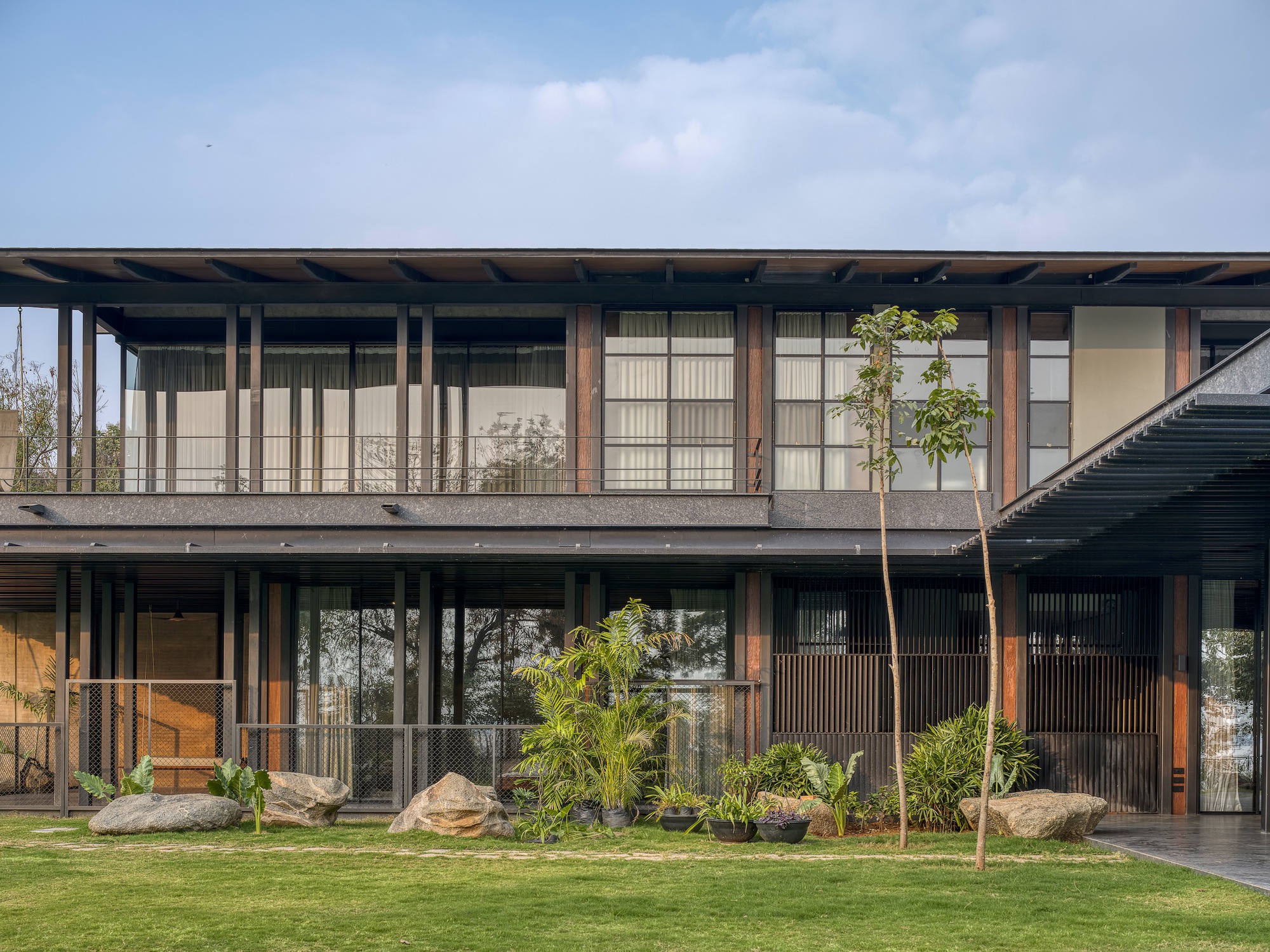 Two-story facade with vertical timber columns and glazing behind a lawn planted with palm and rock outcrops