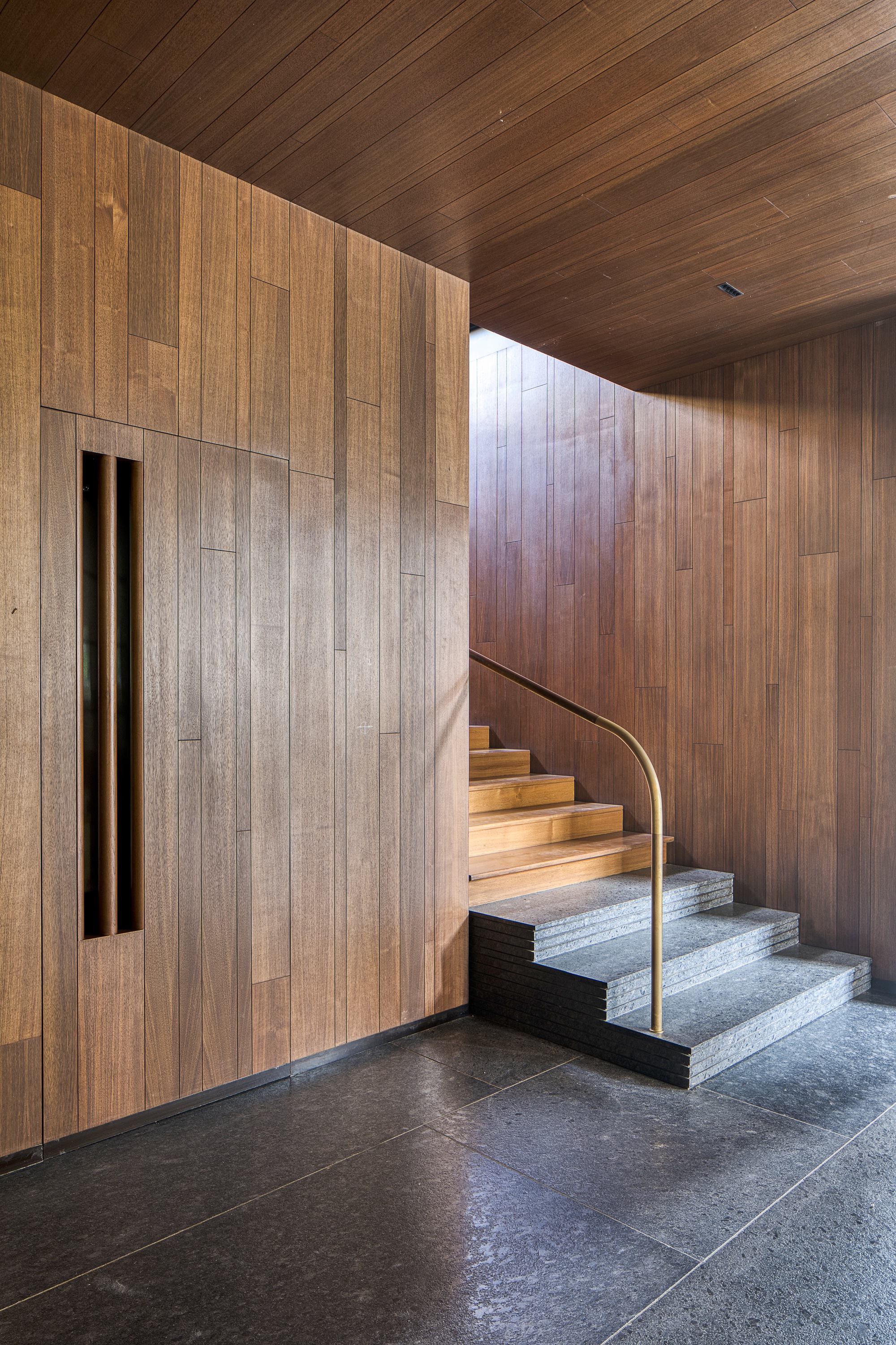 Interior staircase with timber treads and brass rail ascending beside a timber-clad wall with slot window