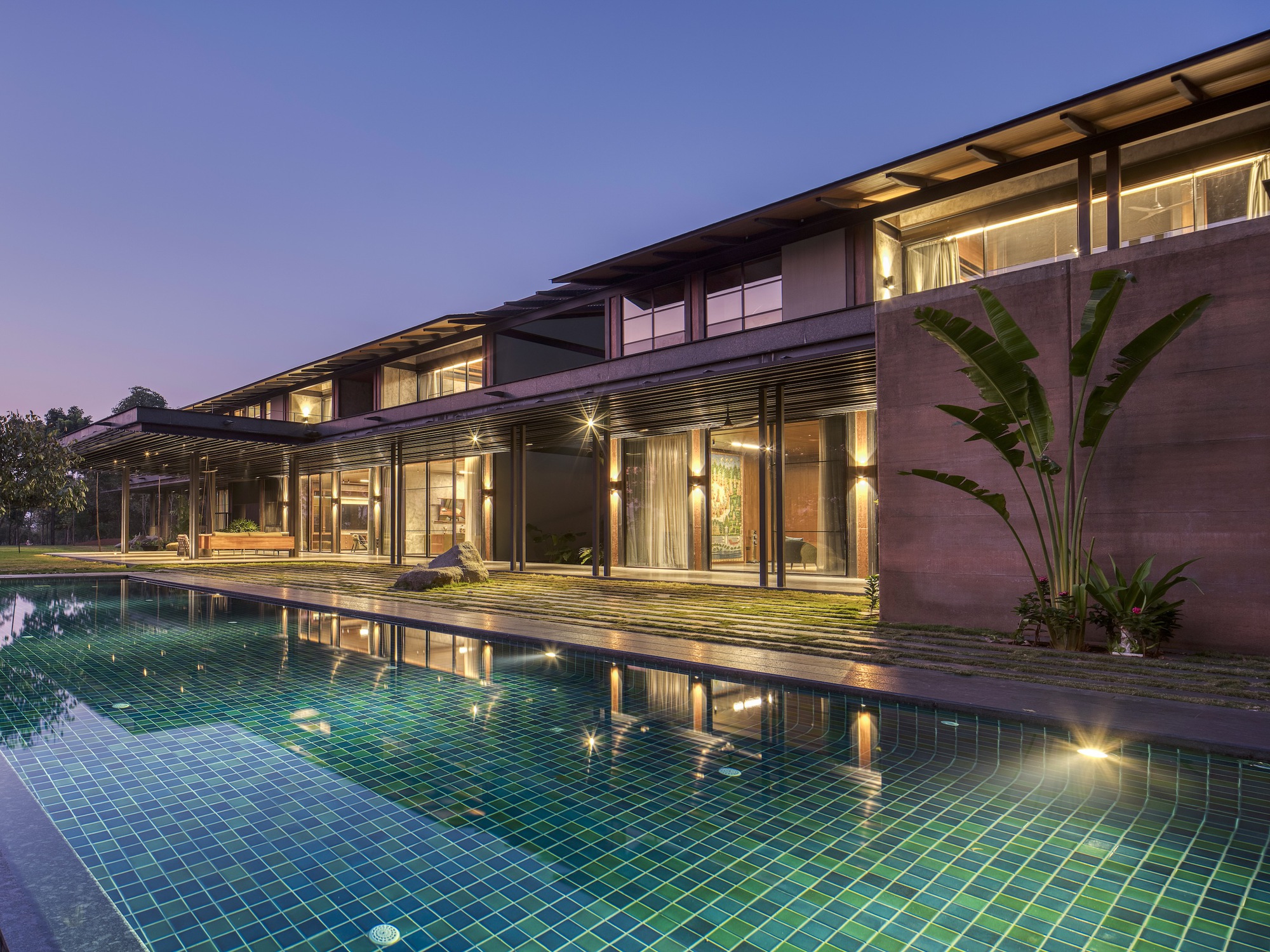 Pool deck with green tile lining reflecting the illuminated interior spaces and timber roof structure at dusk