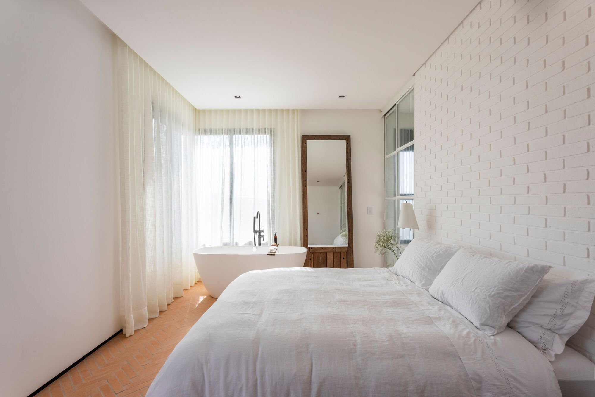 Bedroom with freestanding bathtub beside floor-to-ceiling windows draped in white sheer fabric