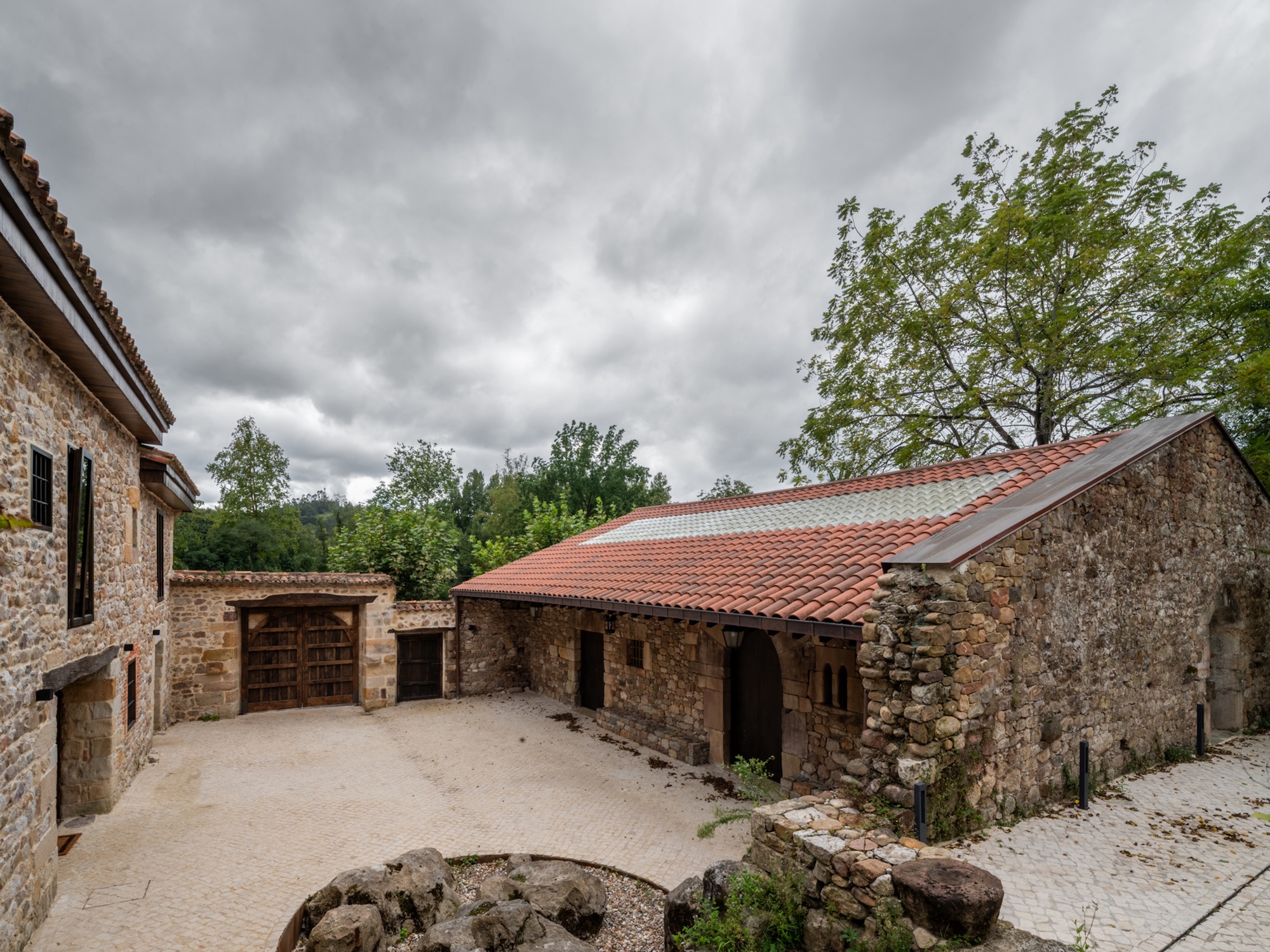 Courtyard view of restored stone buildings with terra cotta roofs surrounded by deciduous trees and cloudy skies